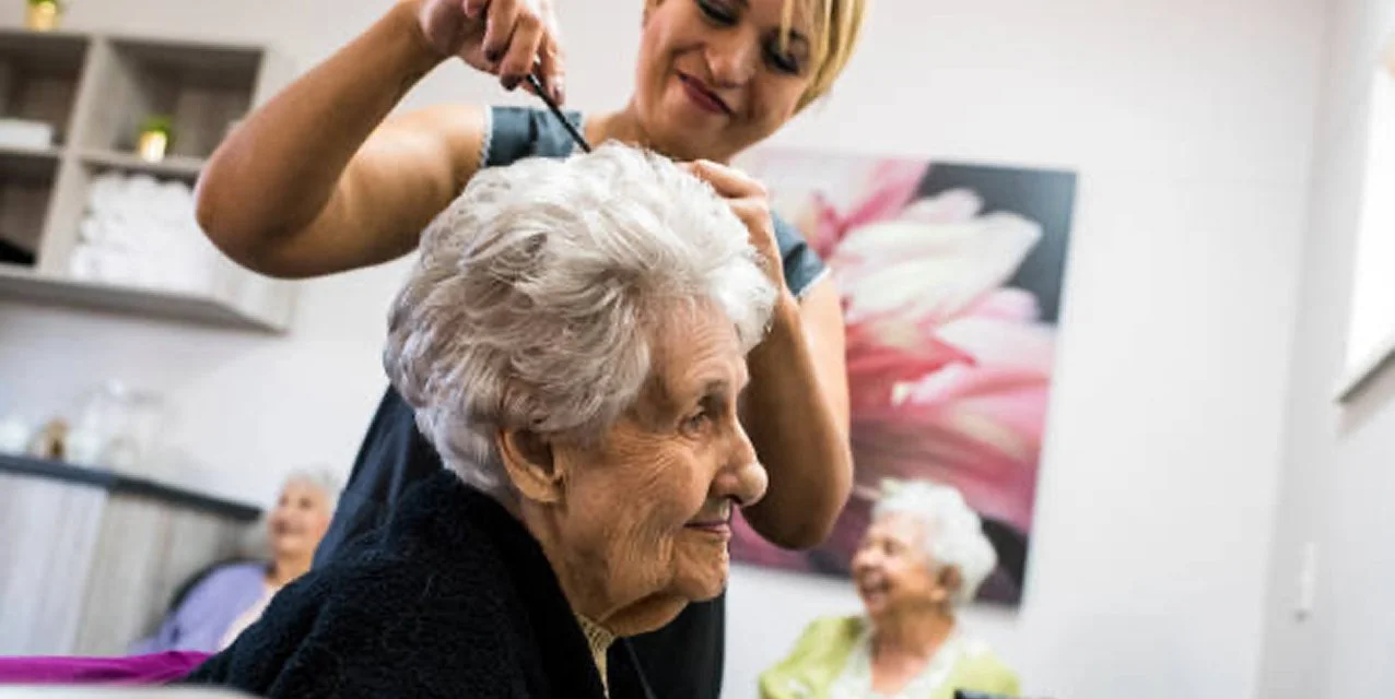 A young woman with short blonde hair giving a haircut to an elderly woman with white hair in a social setting. Two other elderly women are visible in the background, smiling and enjoying the moment.
