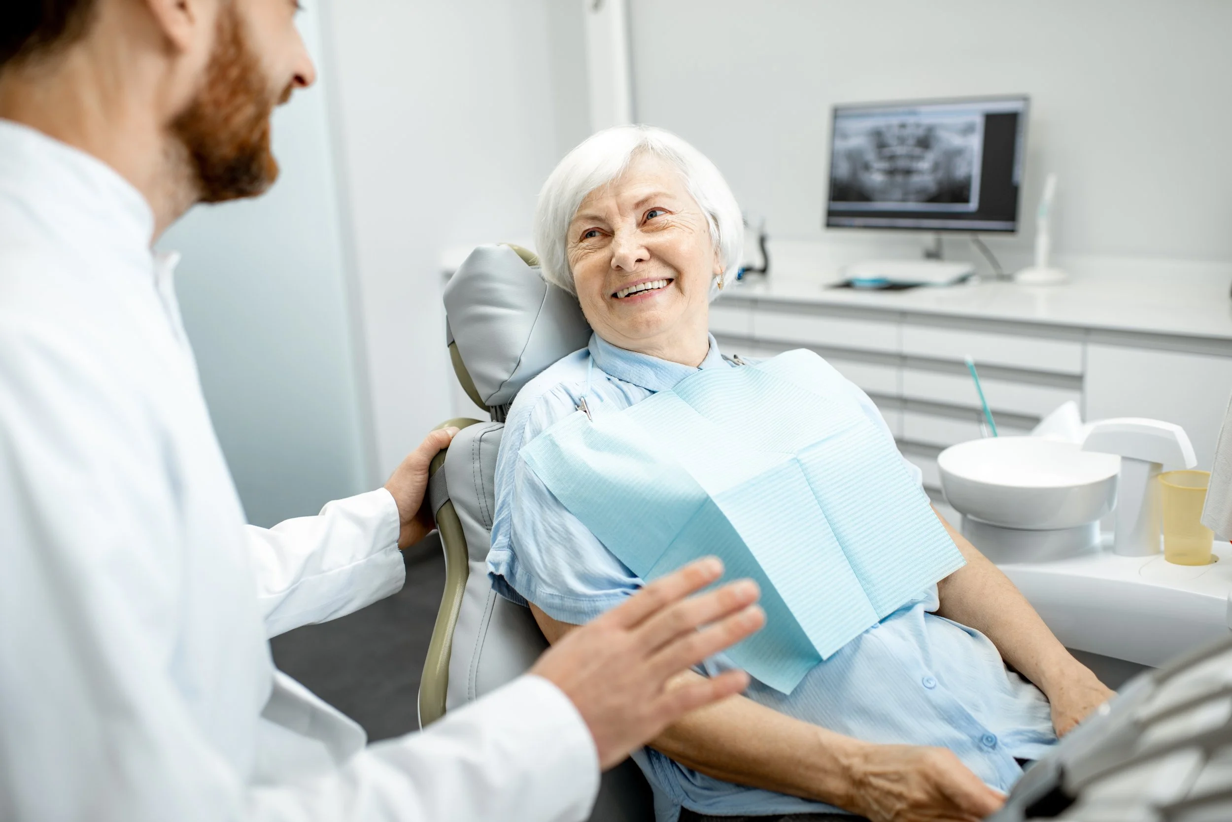 An elderly woman sitting in a dental chair, talking with a dentist in a clinical setting, with dental equipment and a monitor in the background.