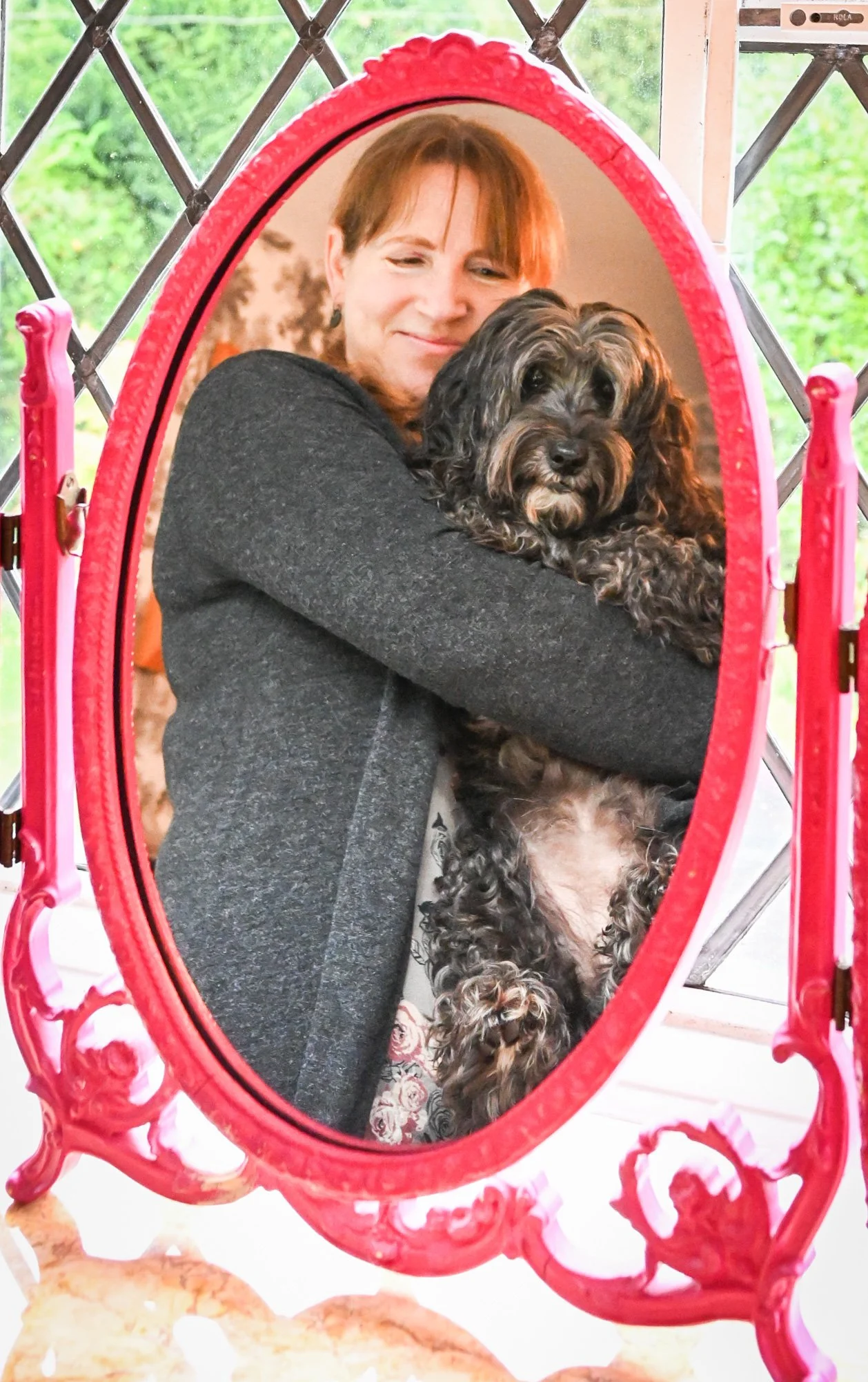 A woman with short brown hair hugging a black and brown curly-haired dog inside a pink oval mirror with a decorative frame, with a green outdoor background visible through a window.