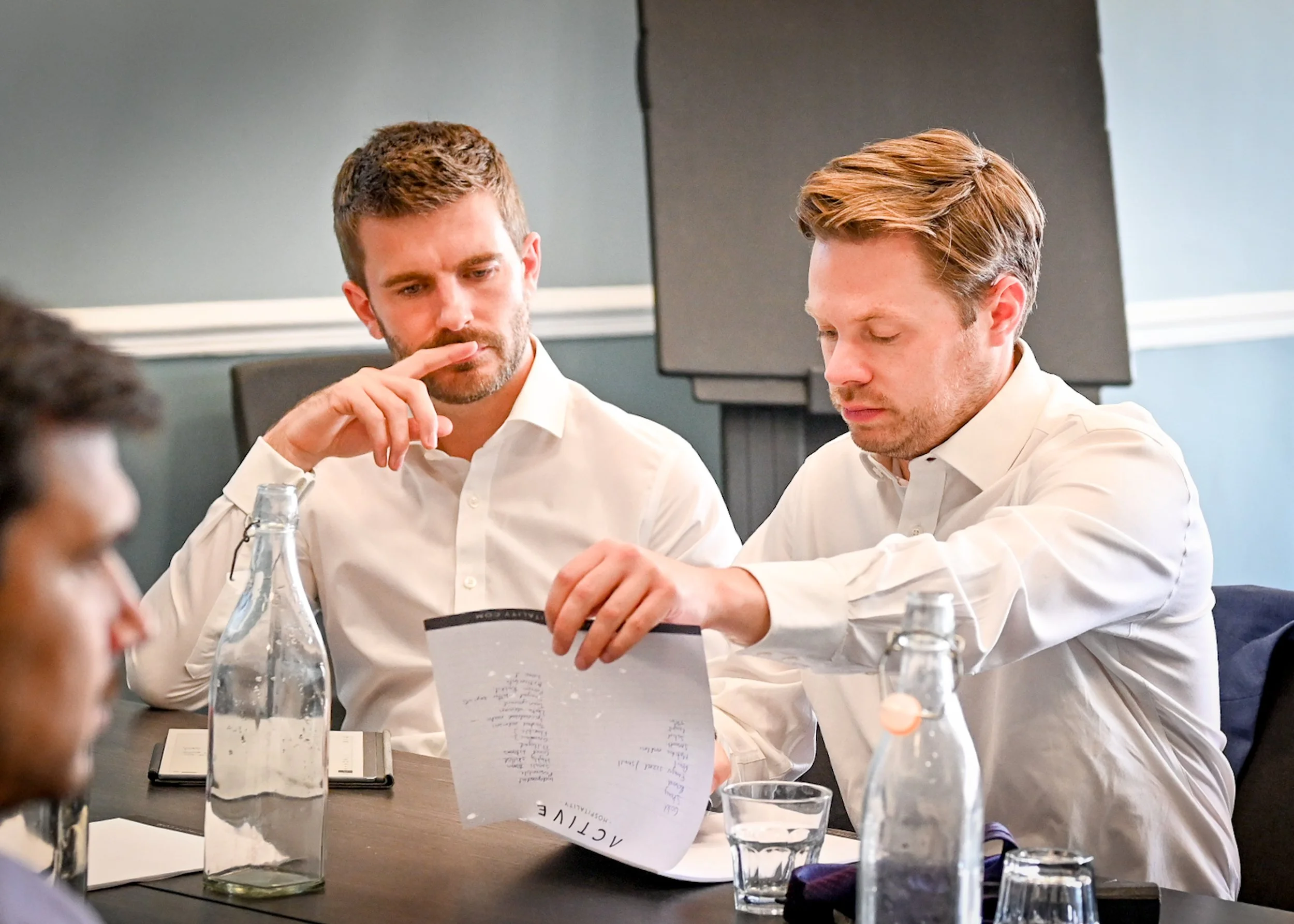 Two men in white shirts sitting at a table, looking at a menu or document, with bottles and glasses on the table.