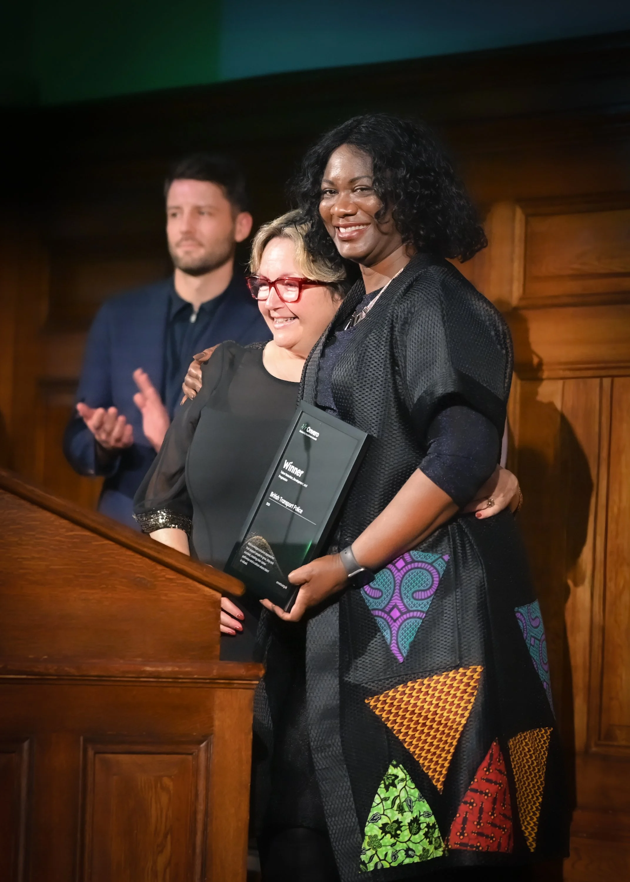 A woman hugging another woman who is holding a trophy or award, while a man claps in the background, at an indoor award ceremony with wooden paneling.