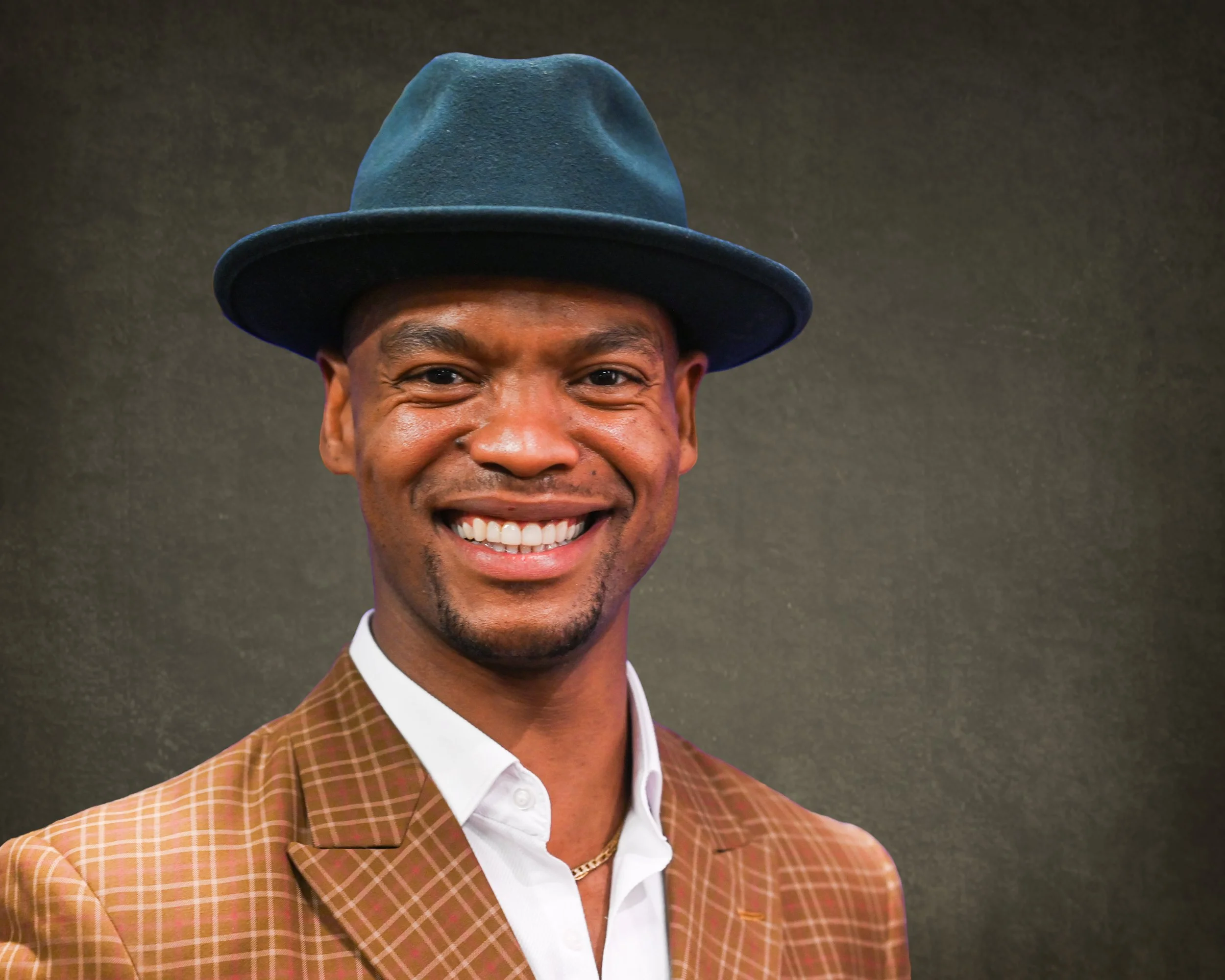 A smiling man wearing a plaid brown blazer, white shirt, gold chain, and a dark blue fedora against a dark background.