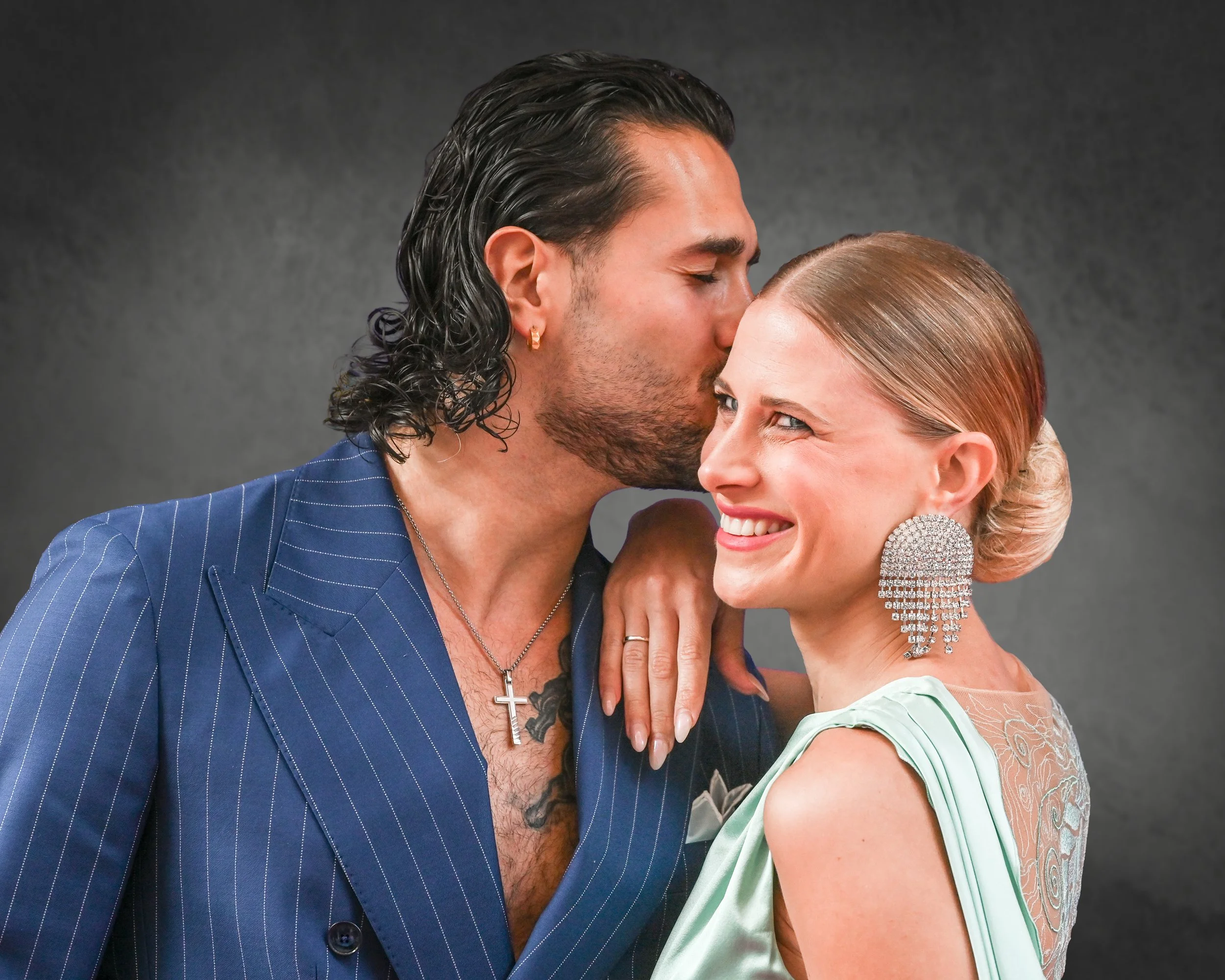 A man with long dark hair, a beard, wearing a blue pinstripe suit, is kissing a woman on the forehead. The woman has light brown hair styled in an updo, is smiling, and wearing a light green dress with lace detailing and large chandelier earrings.