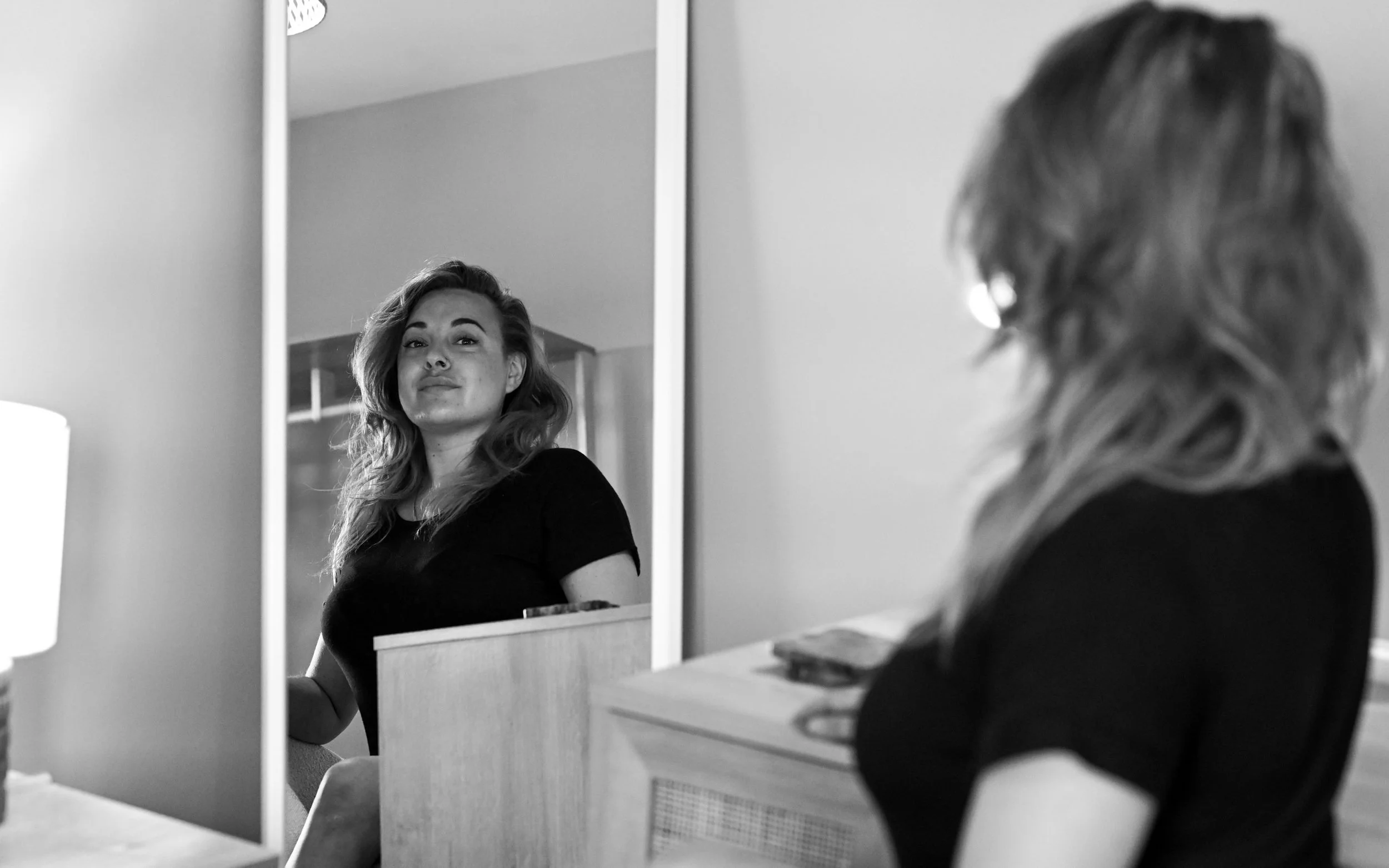 A woman with wavy hair looking at her reflection in a mirror, with her face slightly turned, in a black-and-white photo.