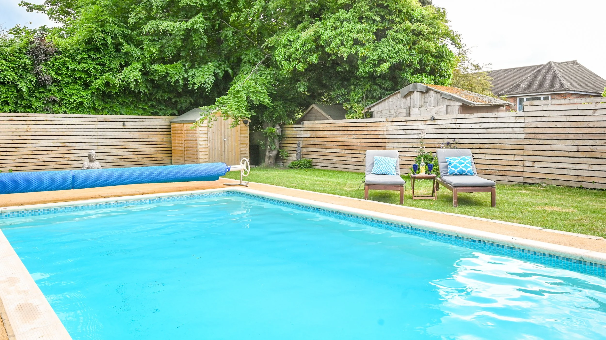 Backyard with a swimming pool surrounded by a wooden fence, two lounge chairs with blue and white pillows, a small side table with two glasses and flowers, green grass, trees, and a small shed.