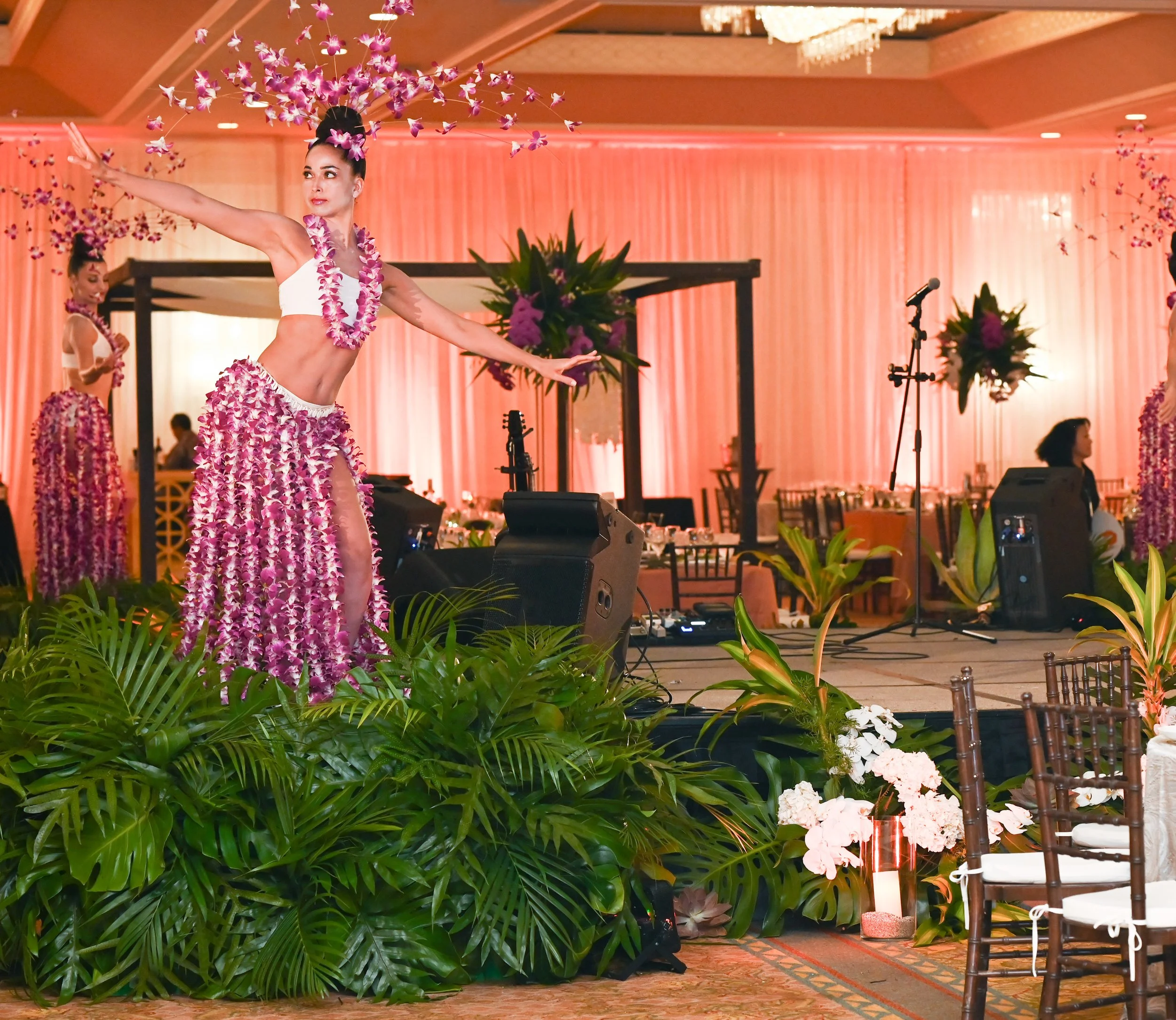 Hula dancers on stage in a decorated event hall with green plants, pink lighting, floral decorations, and tables with chairs.
