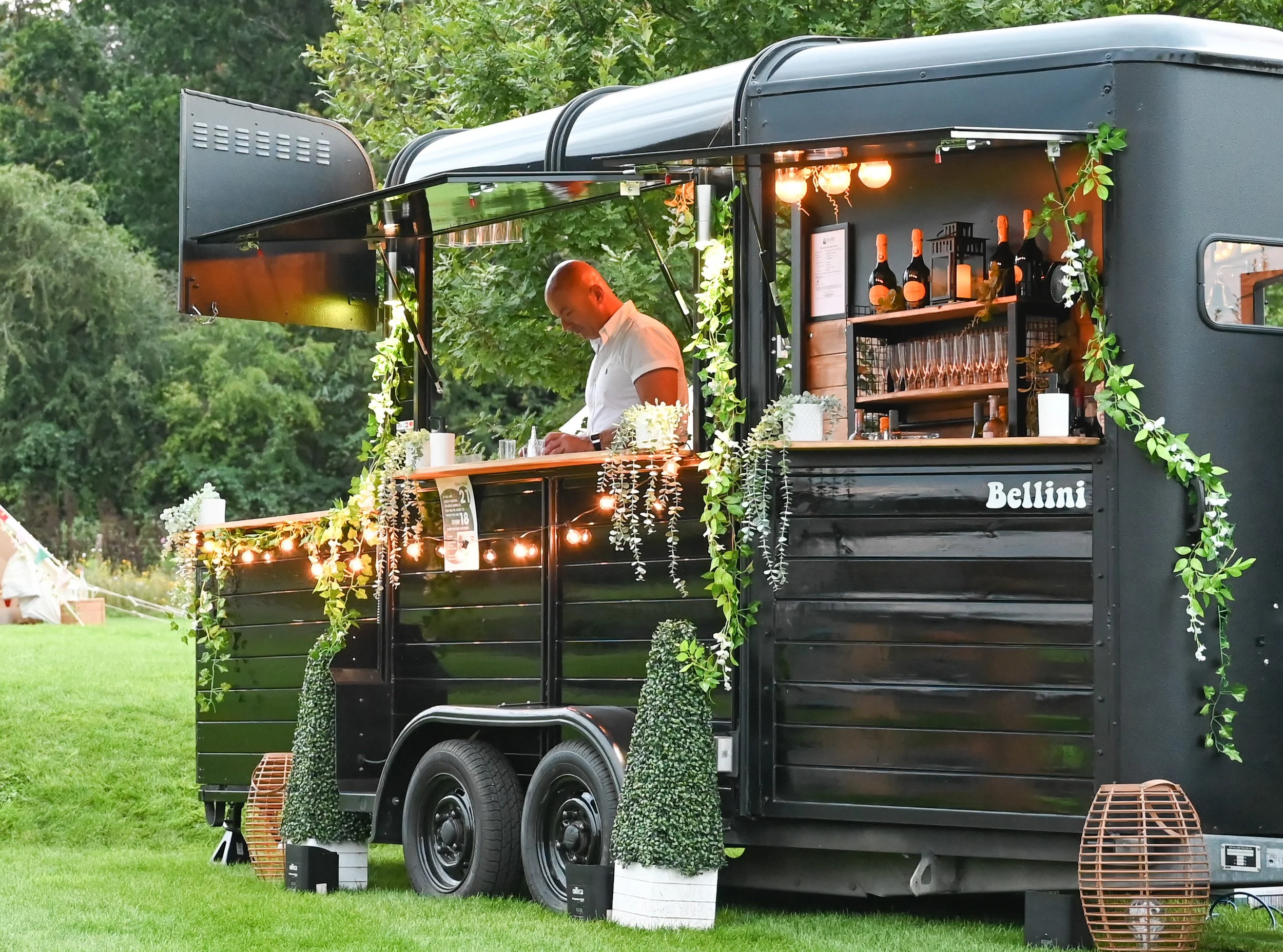 A black food truck decorated with string lights and white flowers, parked on a grassy area with trees in the background. A person is inside the truck, preparing drinks, with wine bottles and glasses displayed behind the counter.
