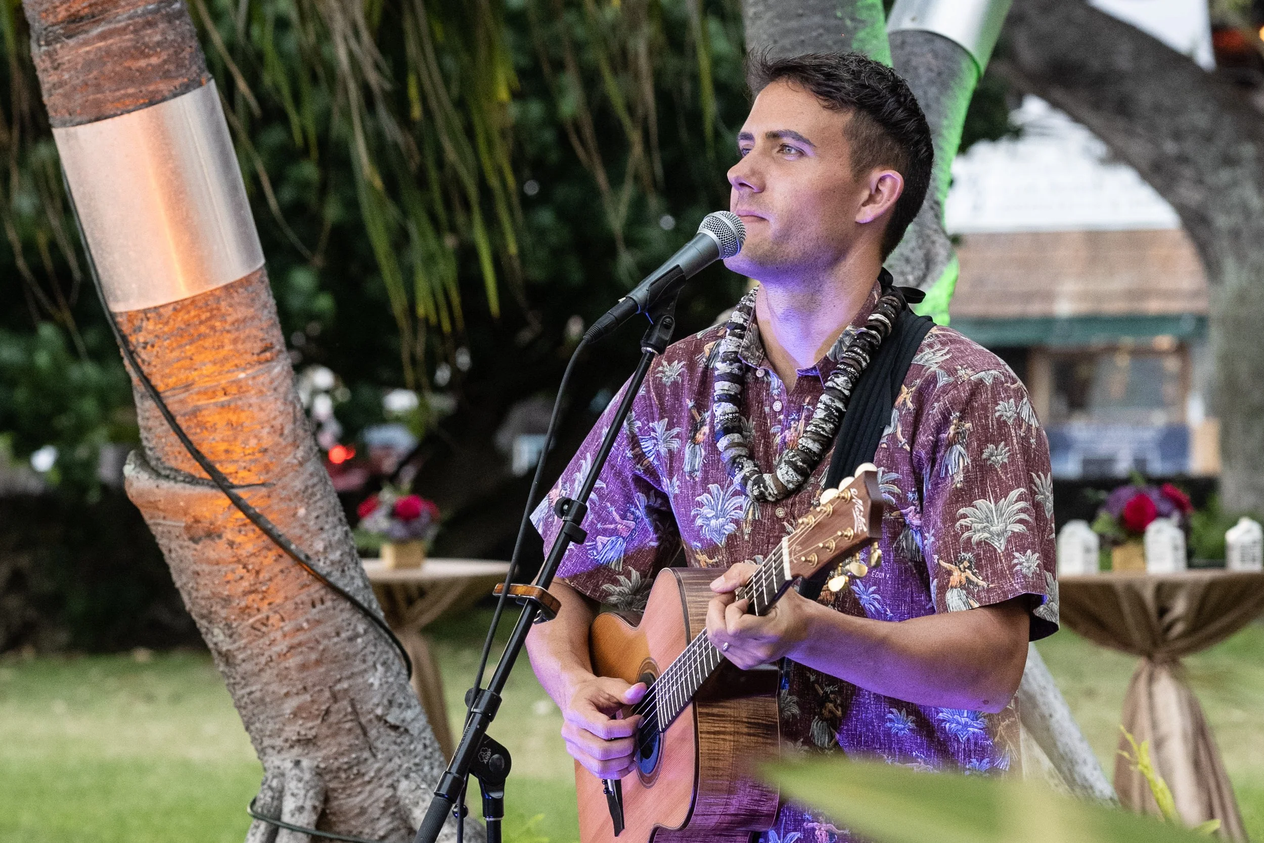 A man wearing a Hawaiian shirt and a shell necklace playing an acoustic guitar and singing into a microphone outdoors with trees and decorative tables in the background.