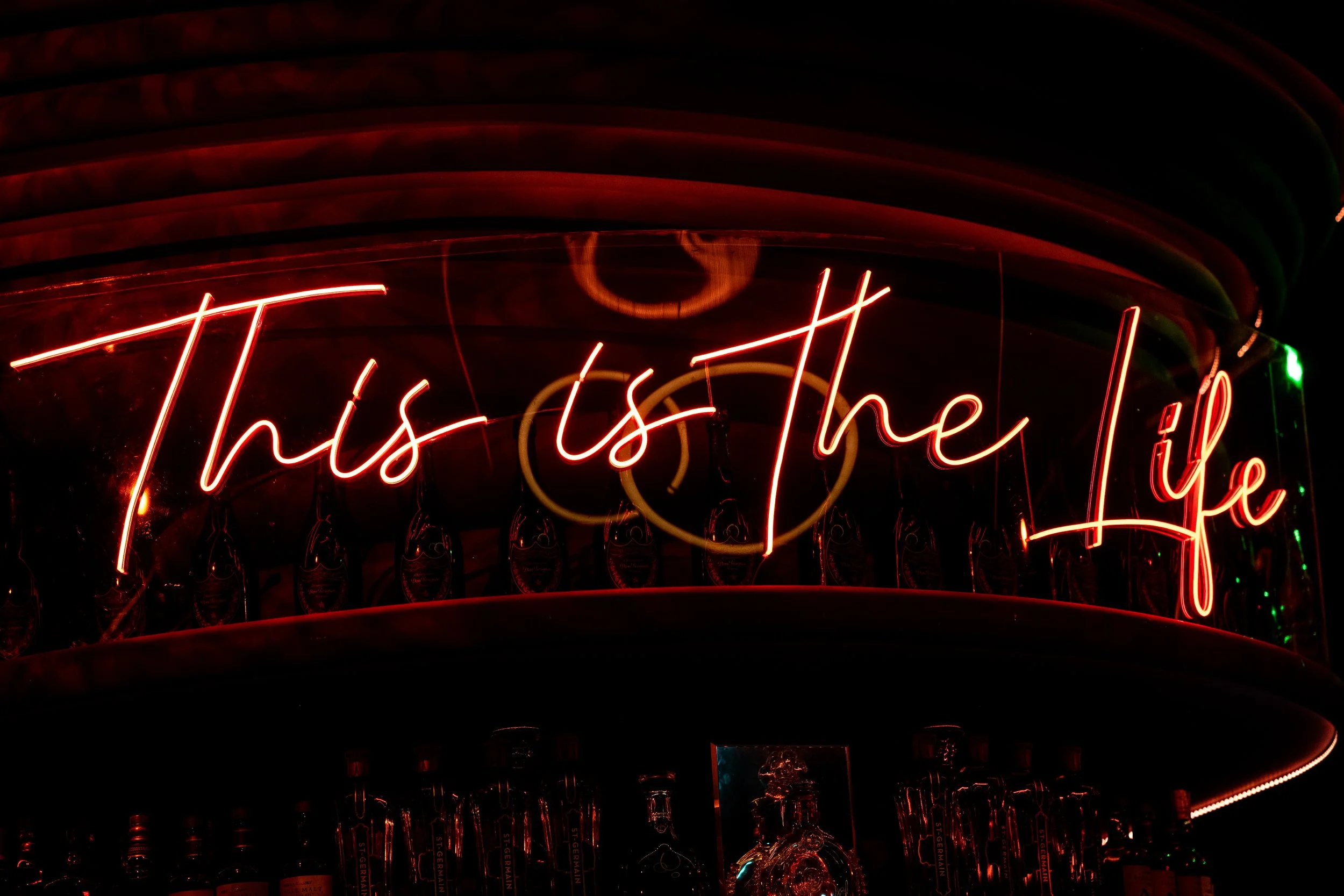 Neon sign that reads 'This is the Life' in red, with bottles displayed below and a dark background.