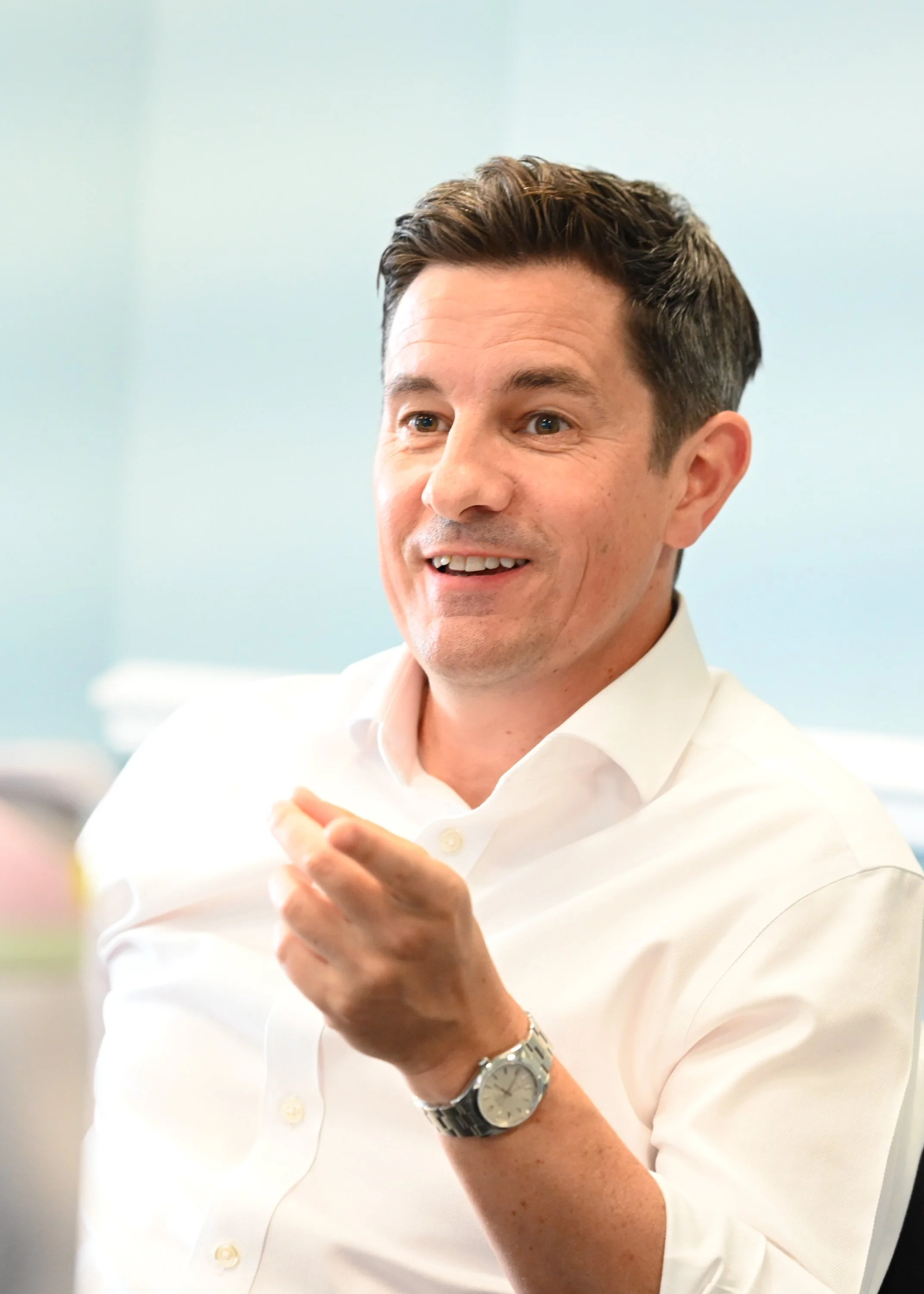 A man smiling and gesturing while wearing a white shirt and a wristwatch in an office setting.