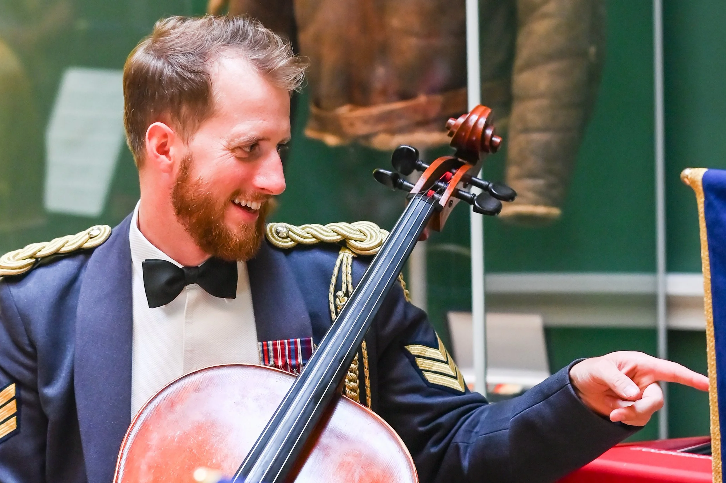 A man dressed in a formal military uniform playing a cello and smiling.