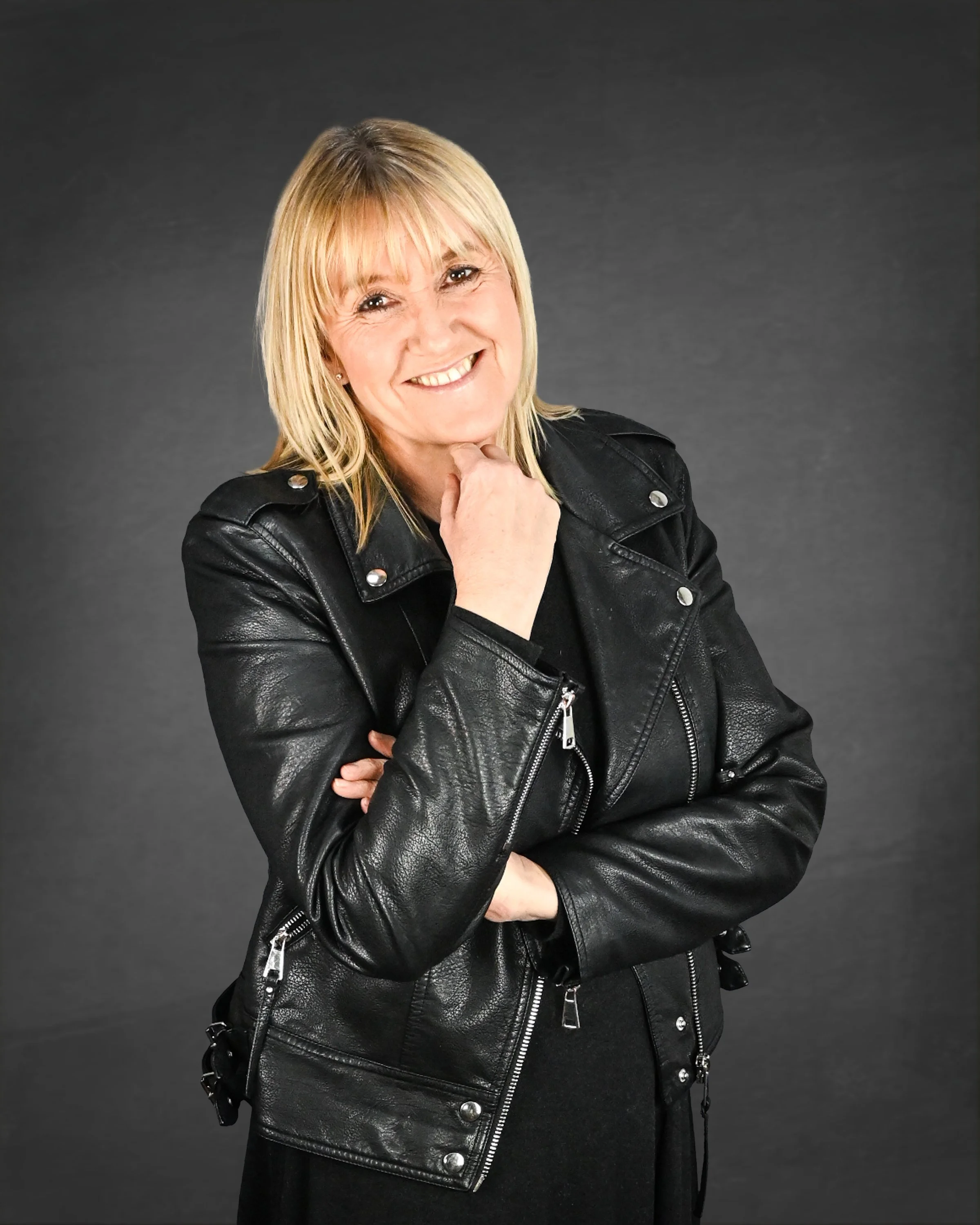 A smiling woman with blonde hair wearing a black leather jacket. She has her left arm crossed and her right hand resting on her chin against a dark grey background.