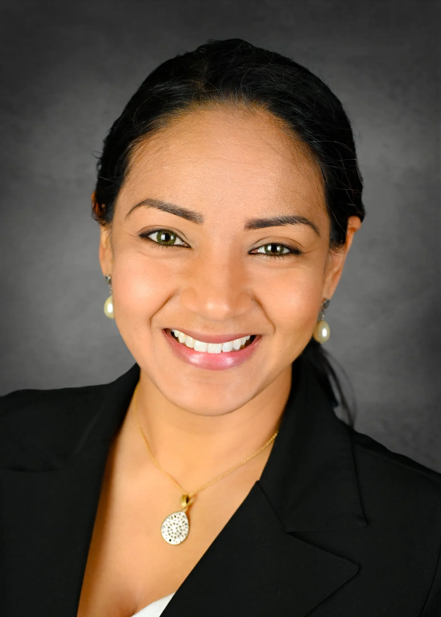 A professional woman with black hair, green eyes, and a bright smile, wearing a black blazer, pearl earrings, and gold jewelry, against a gray background.
