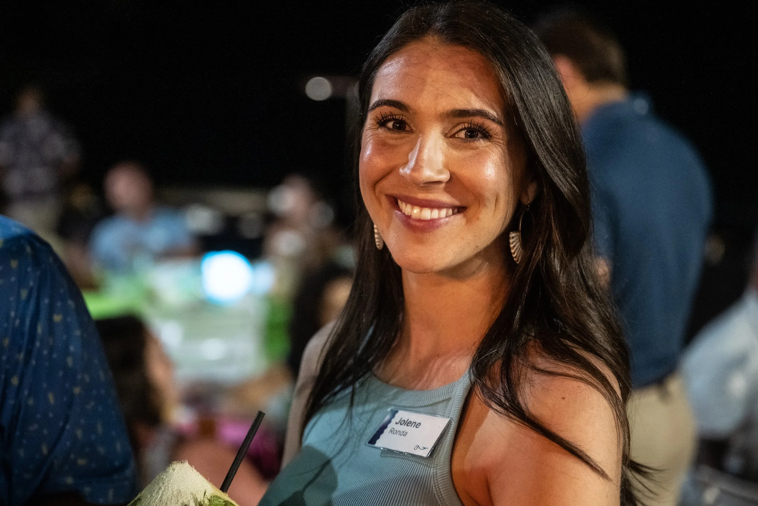 Smiling woman with dark hair, wearing earrings, at a social gathering, holding a drink with a straw, wearing a name tag that reads 'Jolene Ronda'.