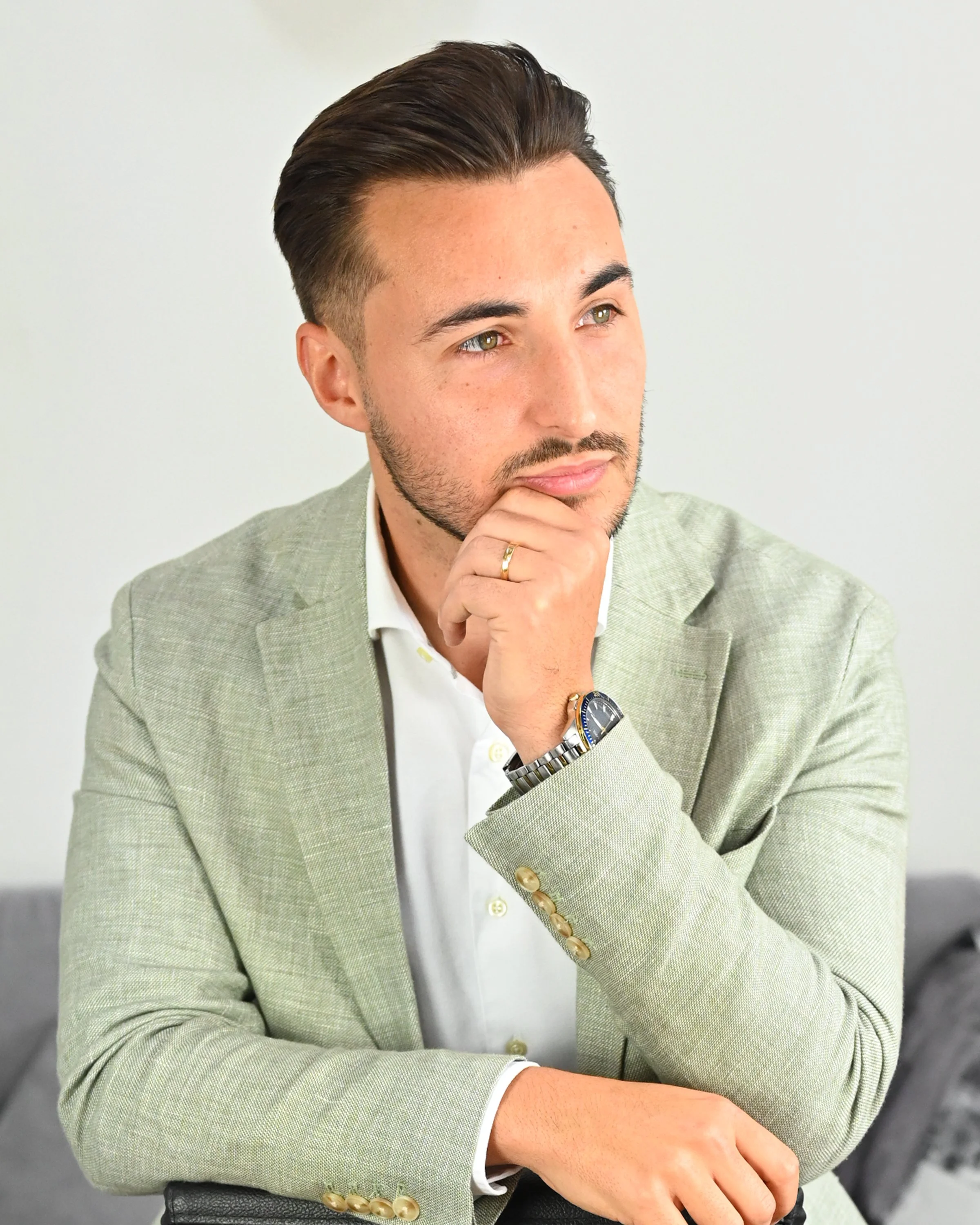 A man with short brown hair and a beard, wearing a light green blazer, white shirt, and a wristwatch, is resting his chin on his hand in a thoughtful pose.