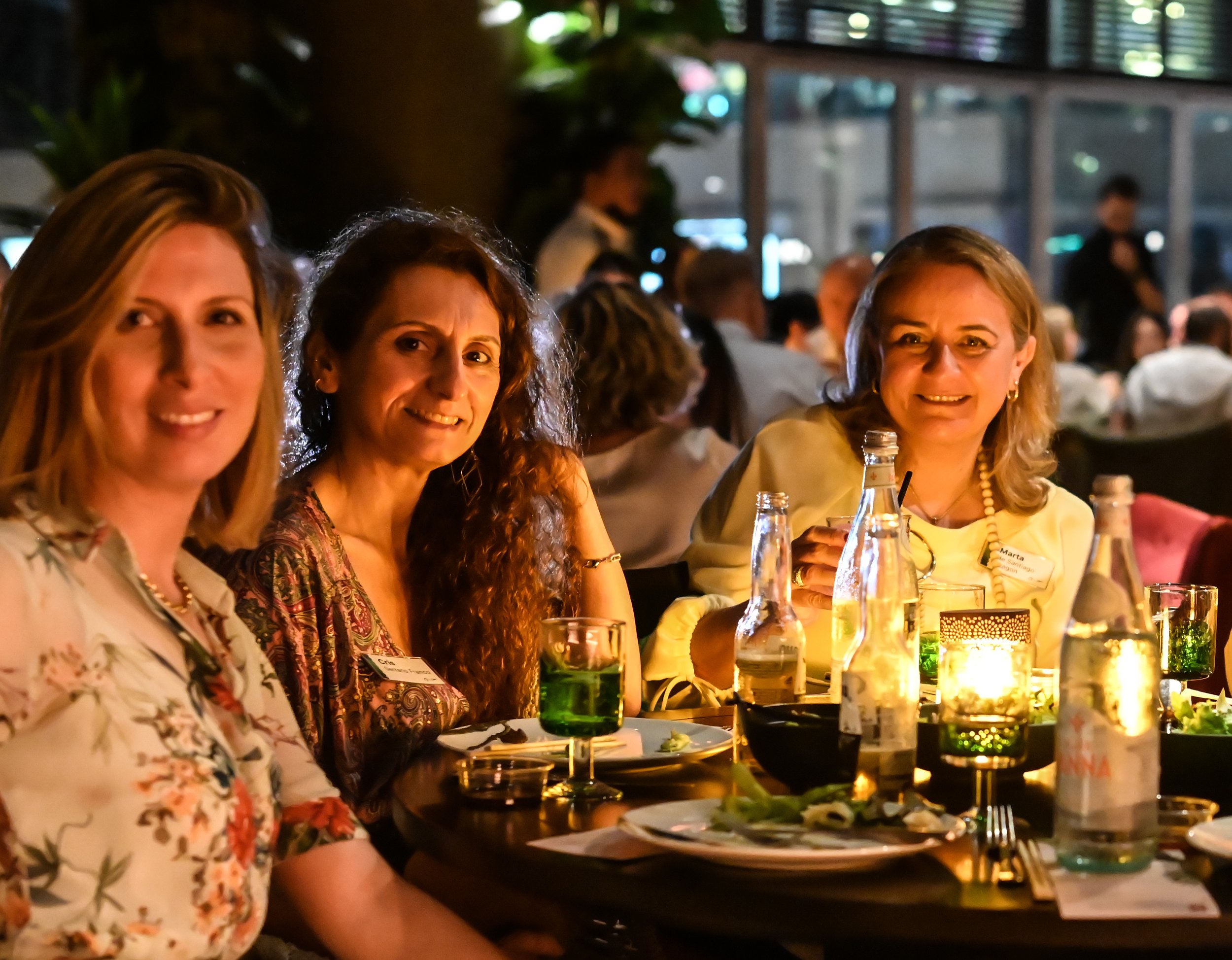 Three women sitting at a restaurant table during an evening event, with plates, drinks, and candlelight.