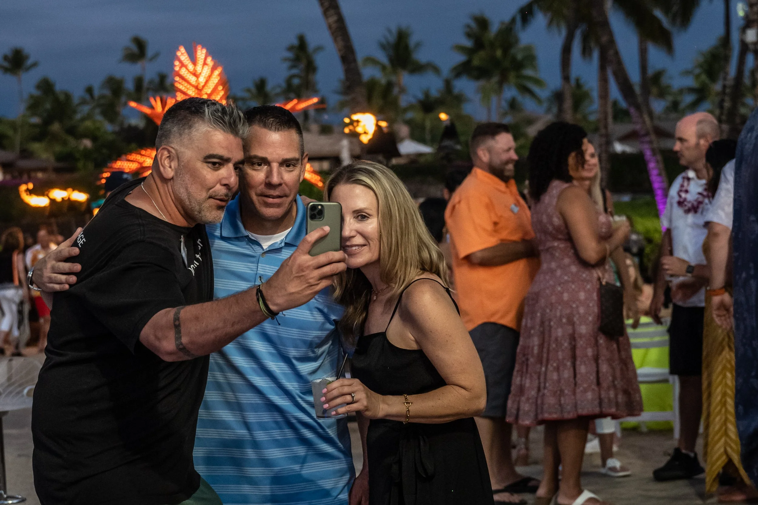 Three people taking a selfie at an outdoor party during the evening with palm trees and lights in the background.