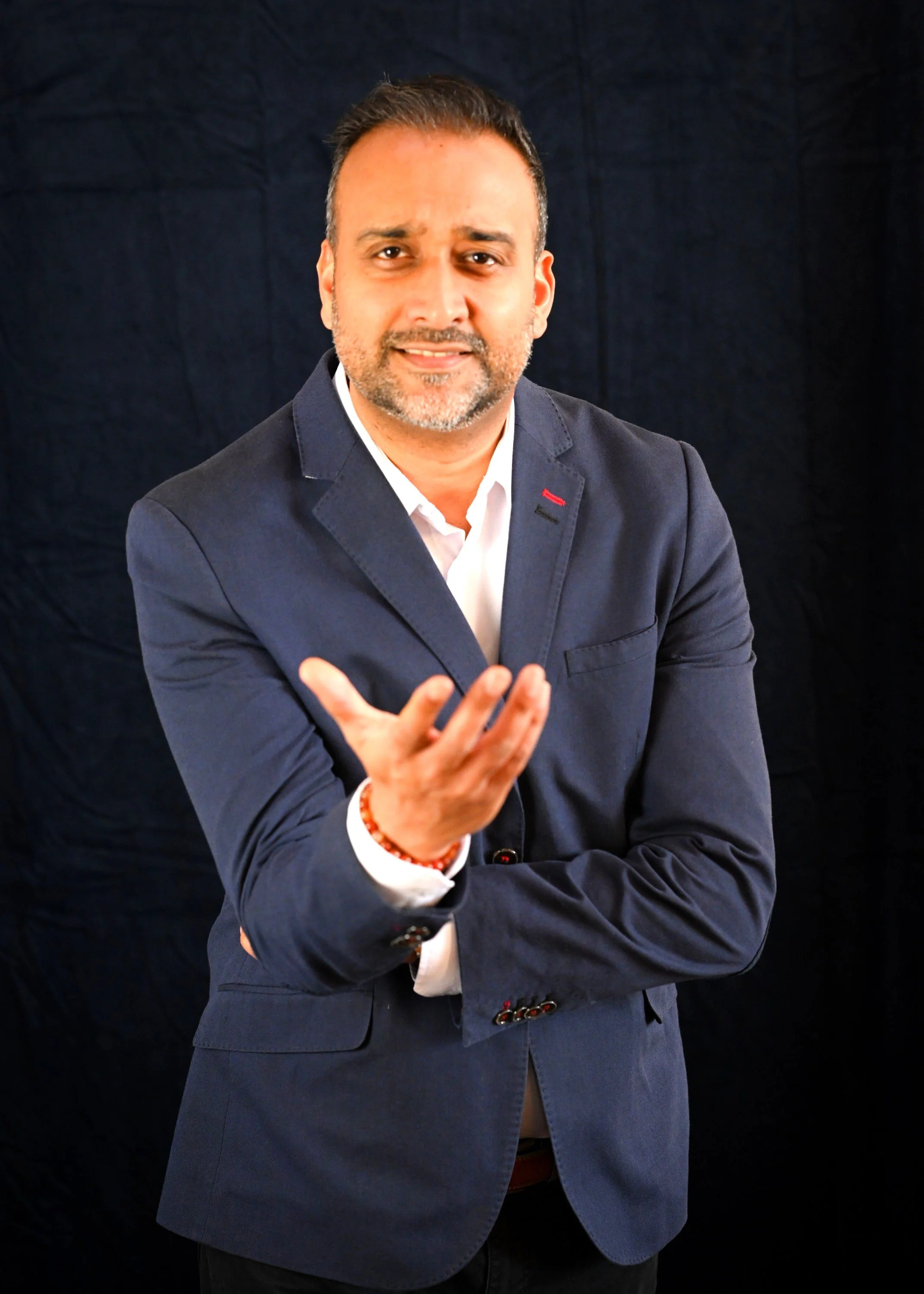 Man in navy blazer and white shirt gesturing towards the camera against a black background.