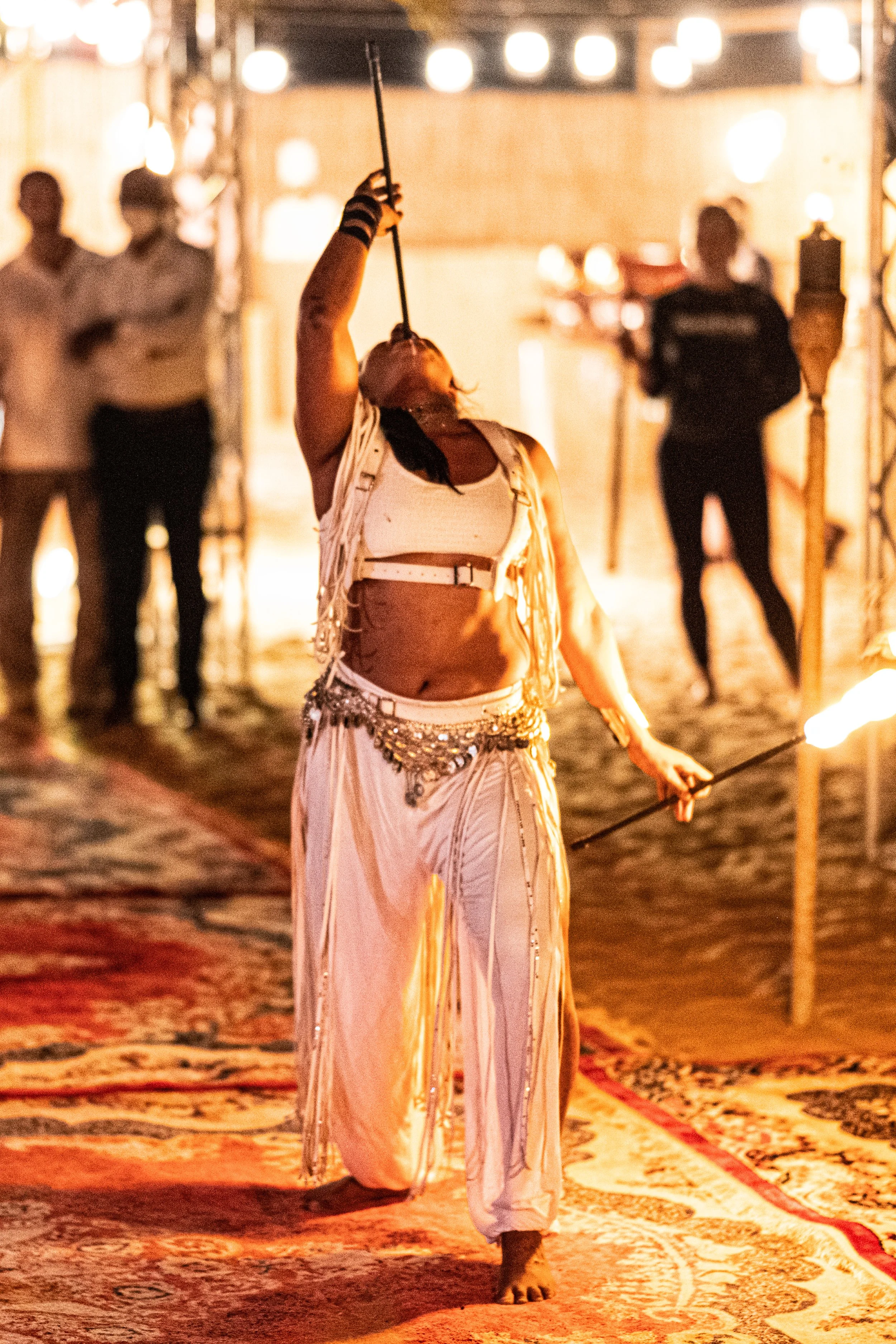 Fire performer spinning fire poi while walking on a patterned carpet, with two people in the background at an indoor event.