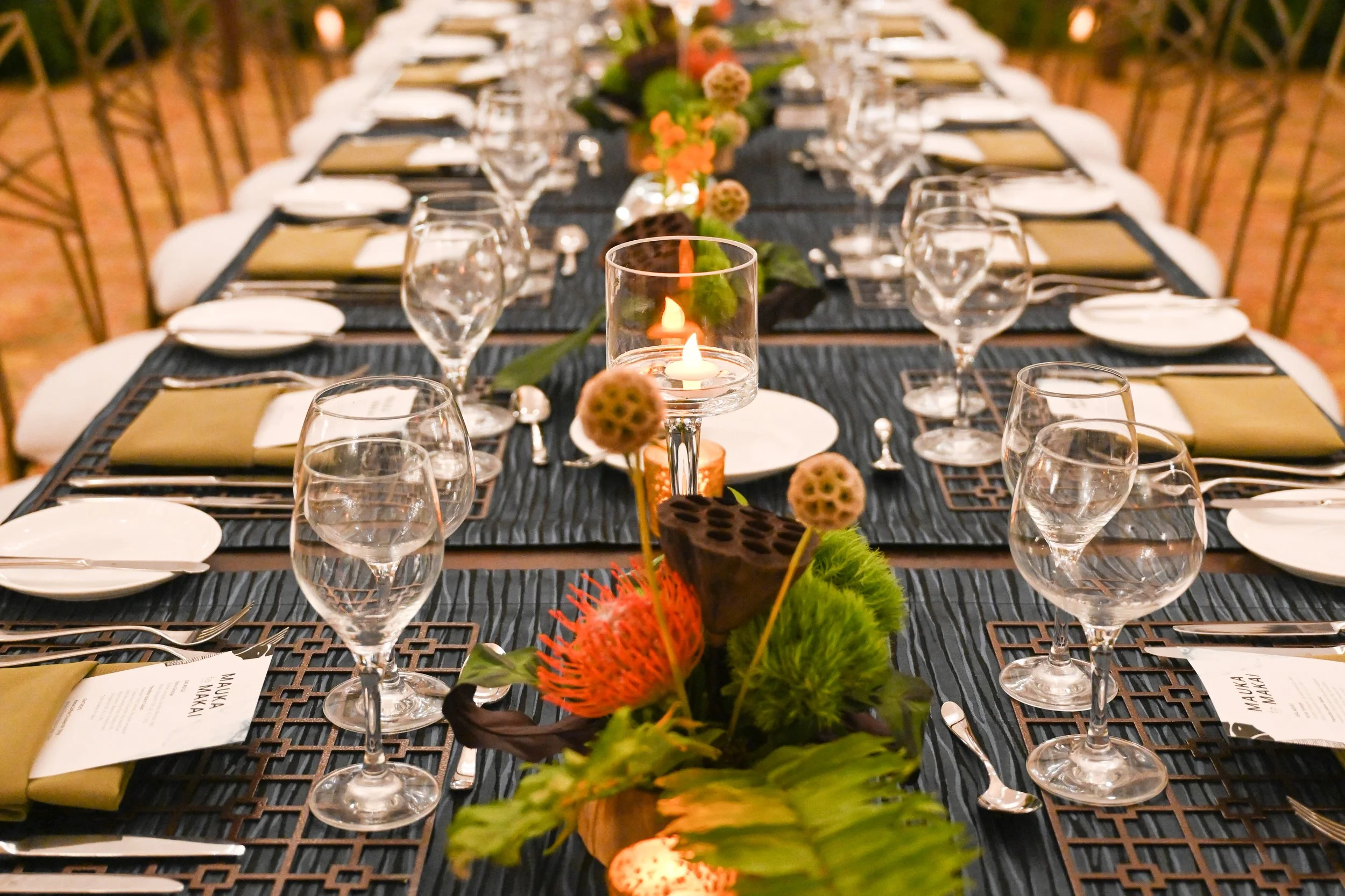 A long banquet table set for a formal event with black tablecloths, white plates, silverware, and multiple wine glasses. A floral centerpiece with tropical flowers and plants runs down the middle, and a candle in a glass holder provides warm lighting