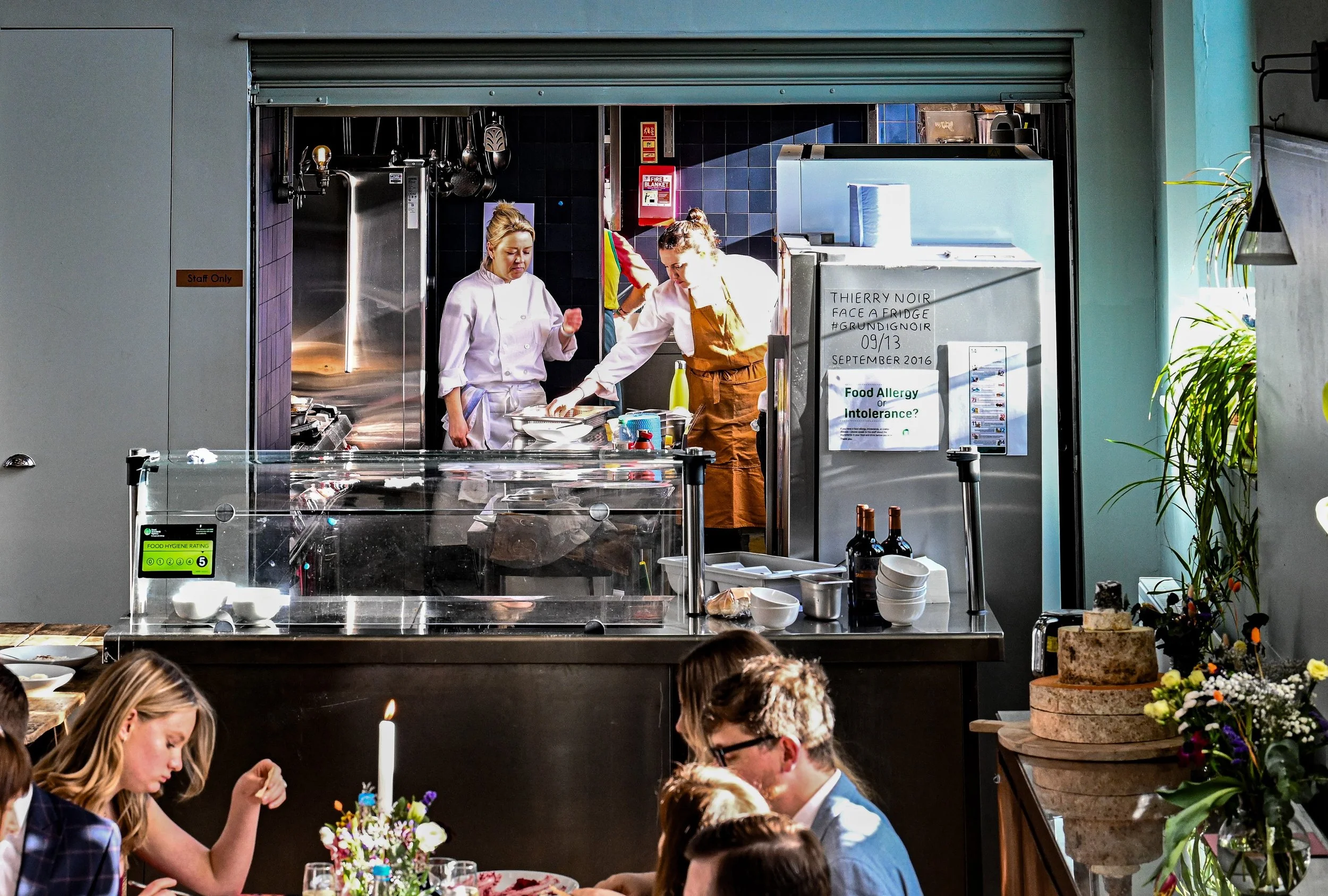 Two chefs in a kitchen preparing food, viewed from a dining area with guests. Decorations include a flower arrangement and a cake on a stand.