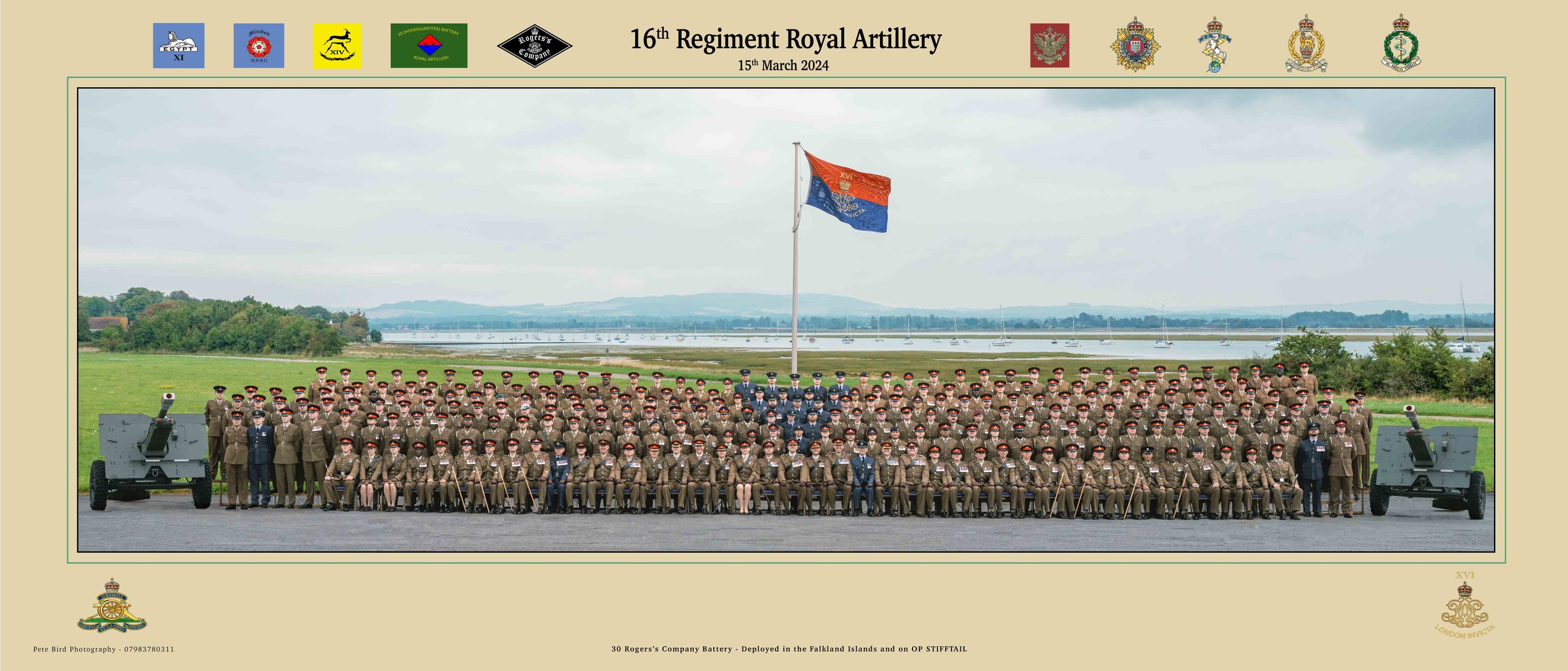 A large group of military personnel in uniform pose together outdoors, with two cannons on either side in front. They are situated on a grassy field with water and boats in the background. A flag is flying on a central pole. The event commemorates th