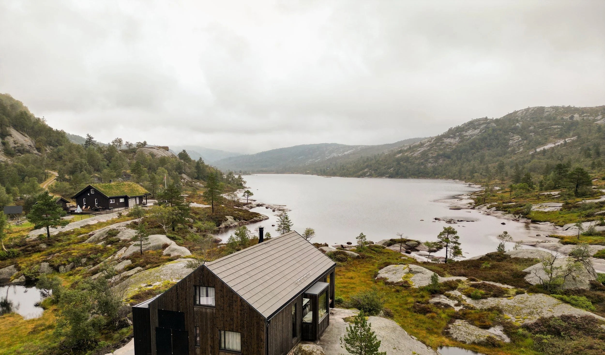 Fjellborg Hytter - Mye grønne trær og busker rundt en innsjø omgitt av fjell på en overskyet dag, med noen svarte hus i landskapet.