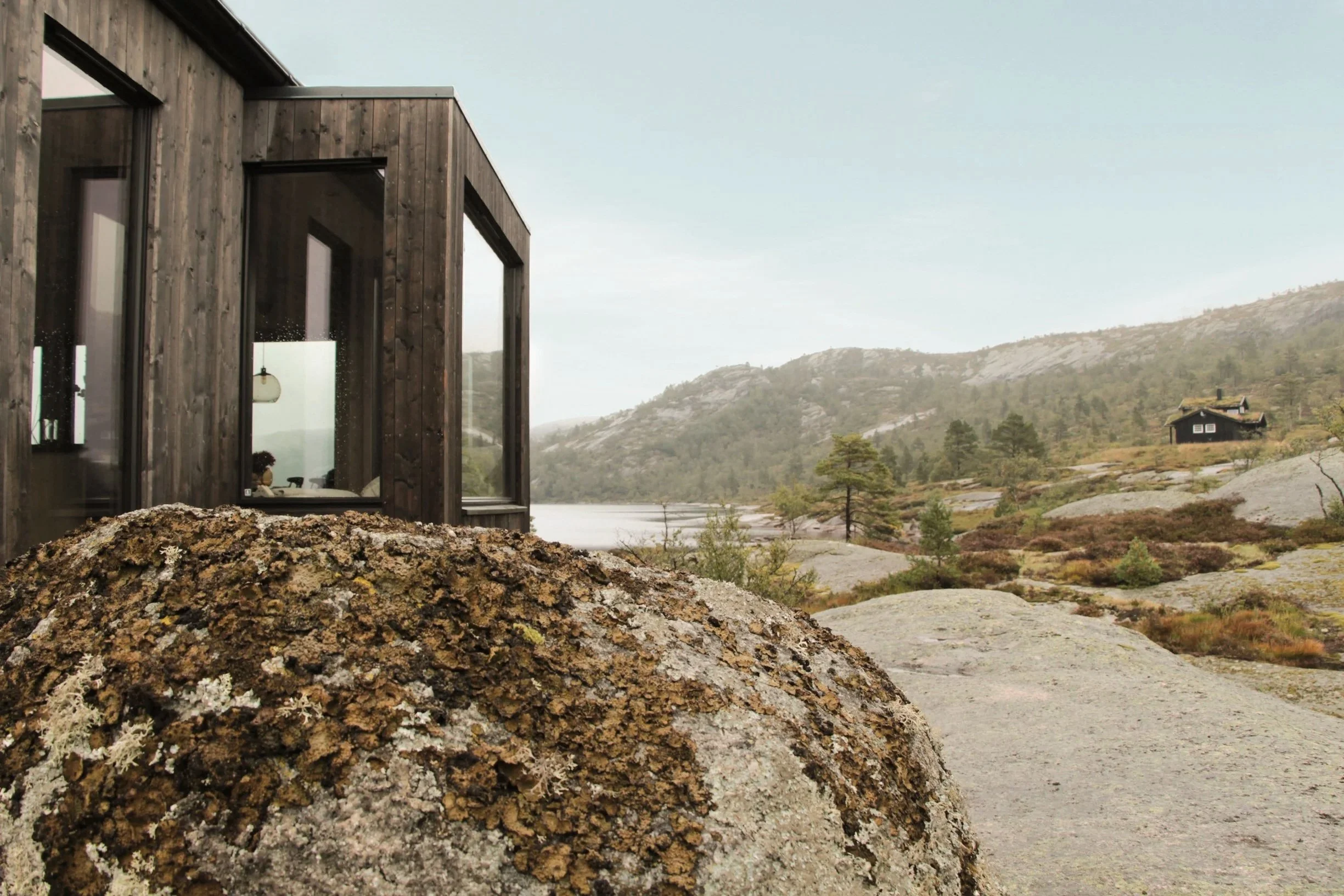 Moderne trehus på fjellandskap med store steiner og spredte trær, fjell og vann i bakgrunnen.