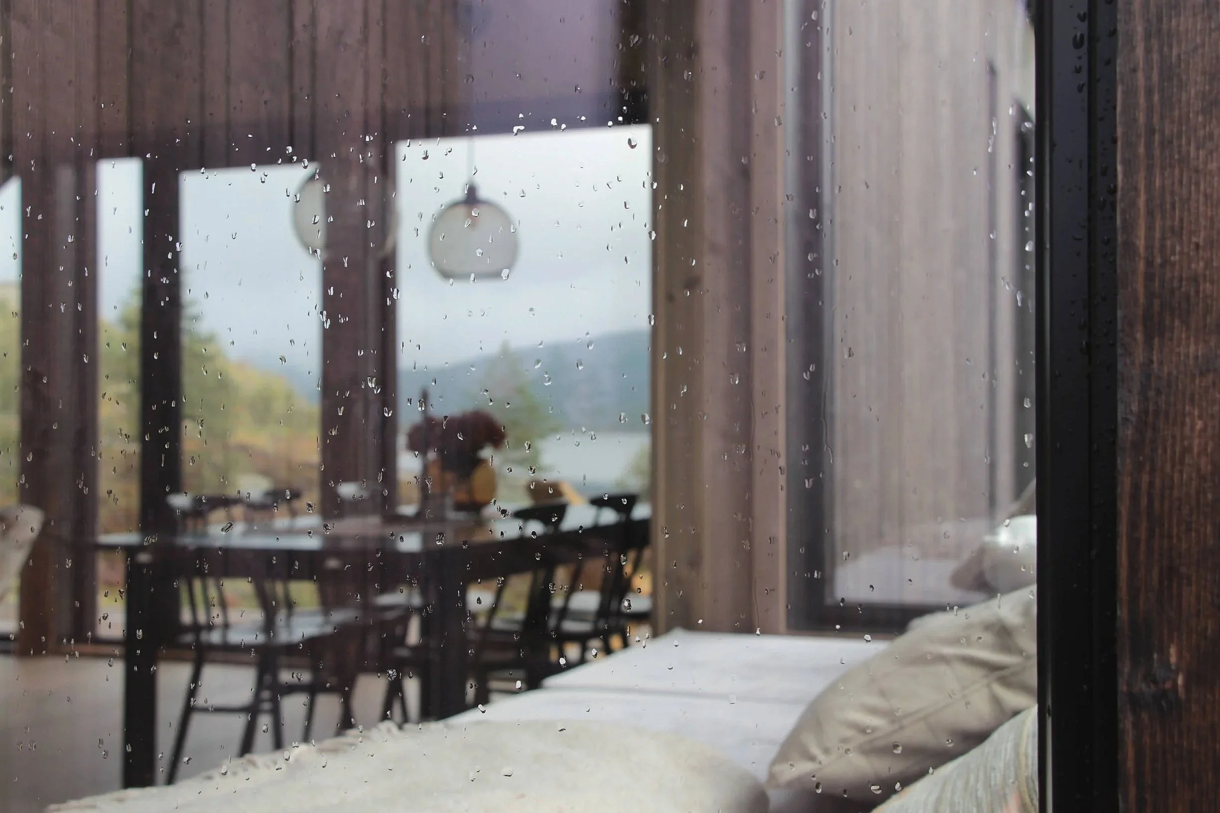 Utsikt inn i et rom sett fra utsiden gjennom et glass med regndråper, med et kjøkken eller spisestue med bord og stoler, og et landskap i bakgrunnen.