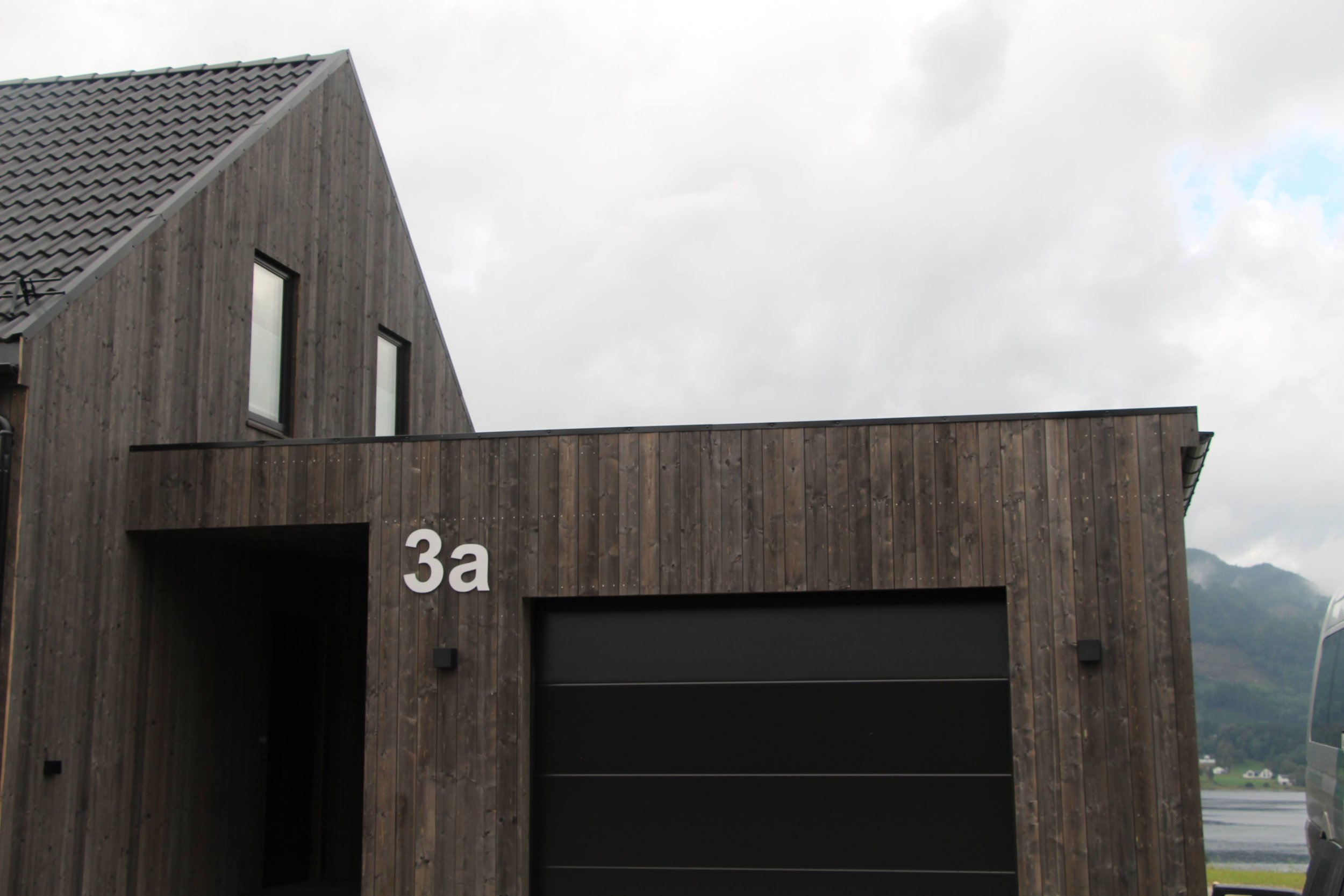 Moderne hus med tre etasjer, kledd i treverk med mørk farge, og en stor garasje. Husnummer 3a ved inngangen, omgitt av landskap med fjell og vann i bakgrunnen under overskyet vær.