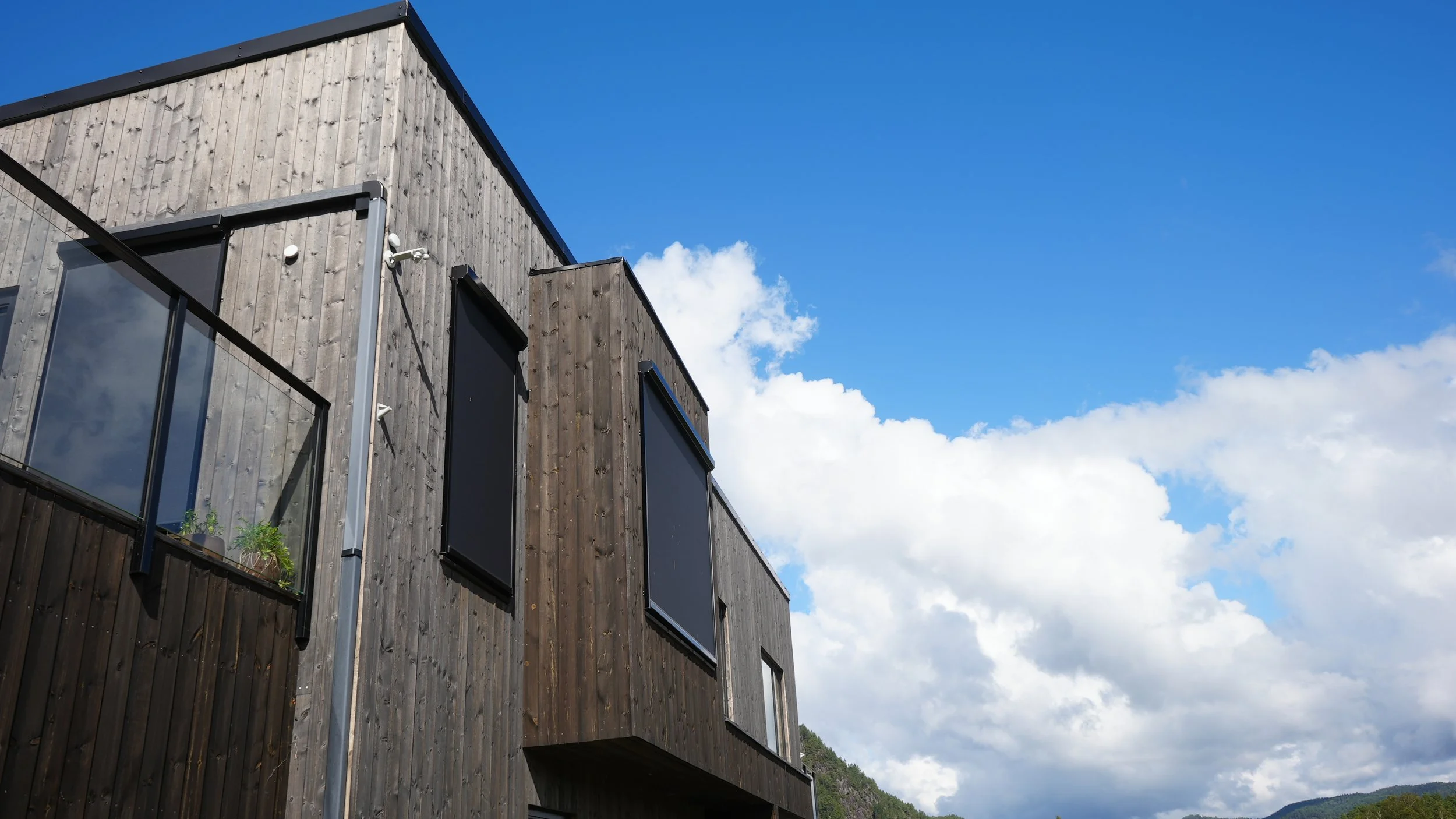 Modern multi-story house with wooden exterior, large windows, and a balcony, under a bright blue sky with white clouds.