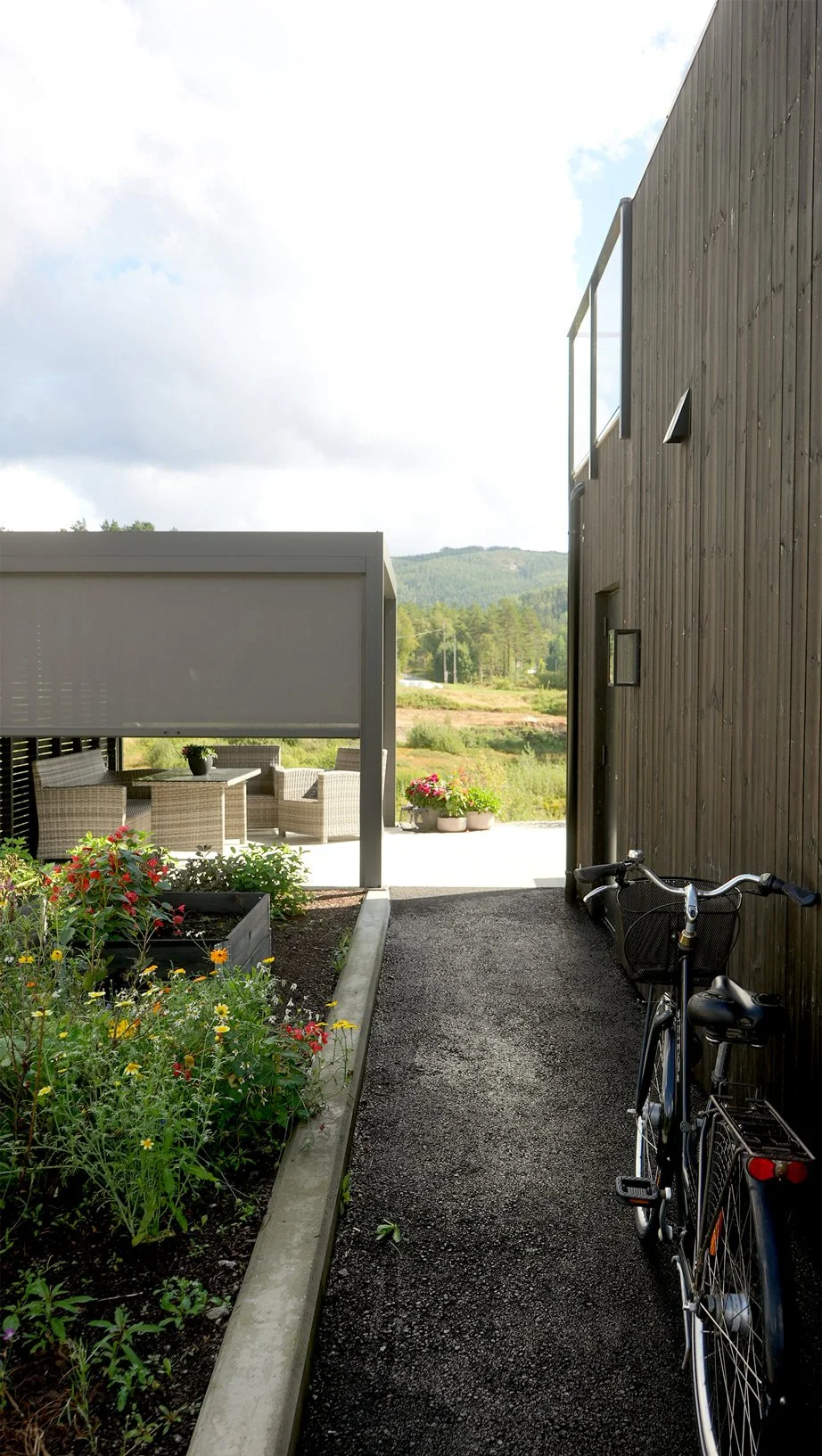 Utsikt mot hage med blomsterbed, terrasse med sittegruppe, sykler parkert langs en vegg, mot fjell og skyet himmel i bakgrunnen.