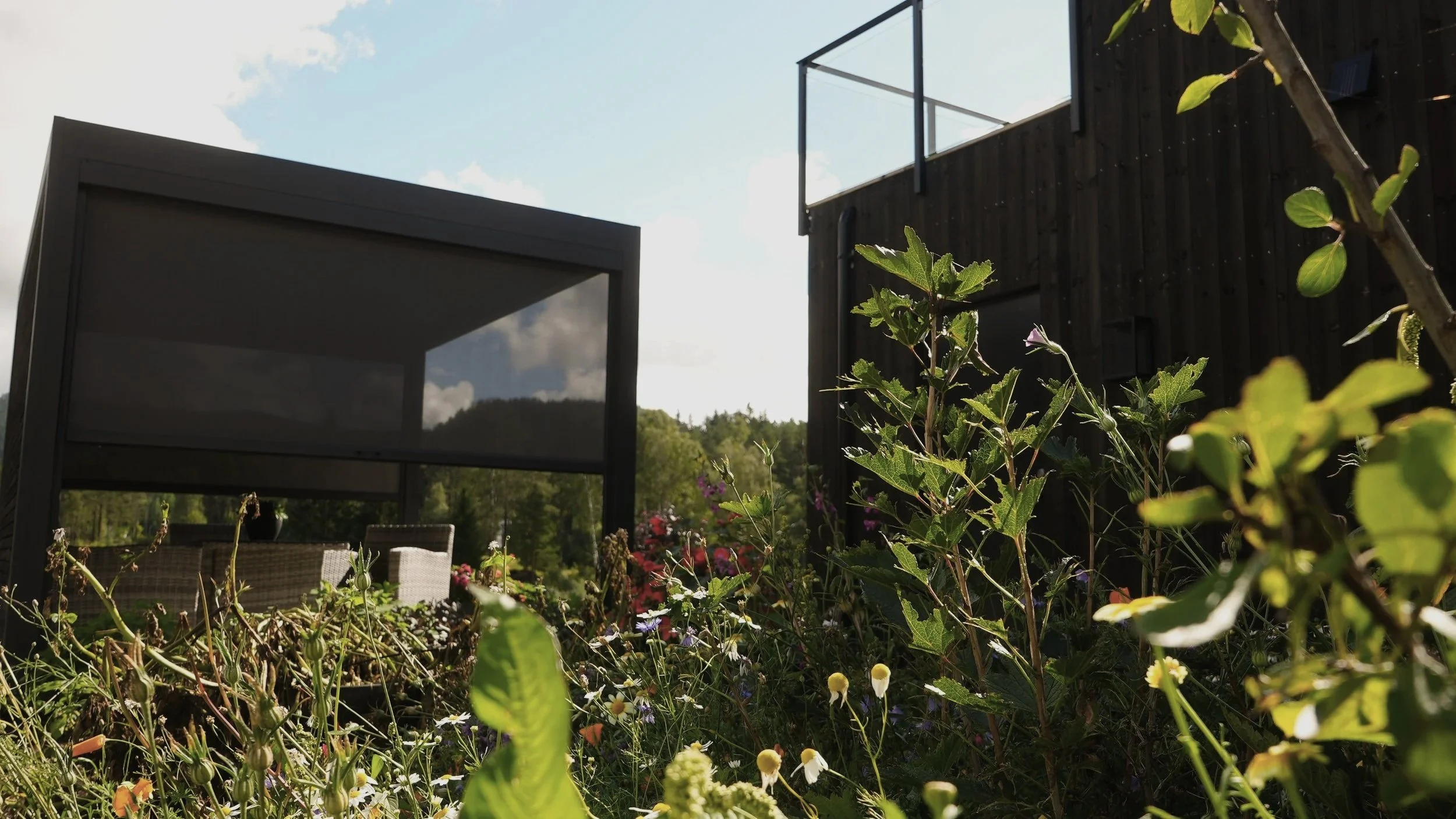 Uteplass med moderne møbler omgitt av blomster og grønne planter, med et svart hus i bakgrunnen og en blå himmel med noen skyer.
