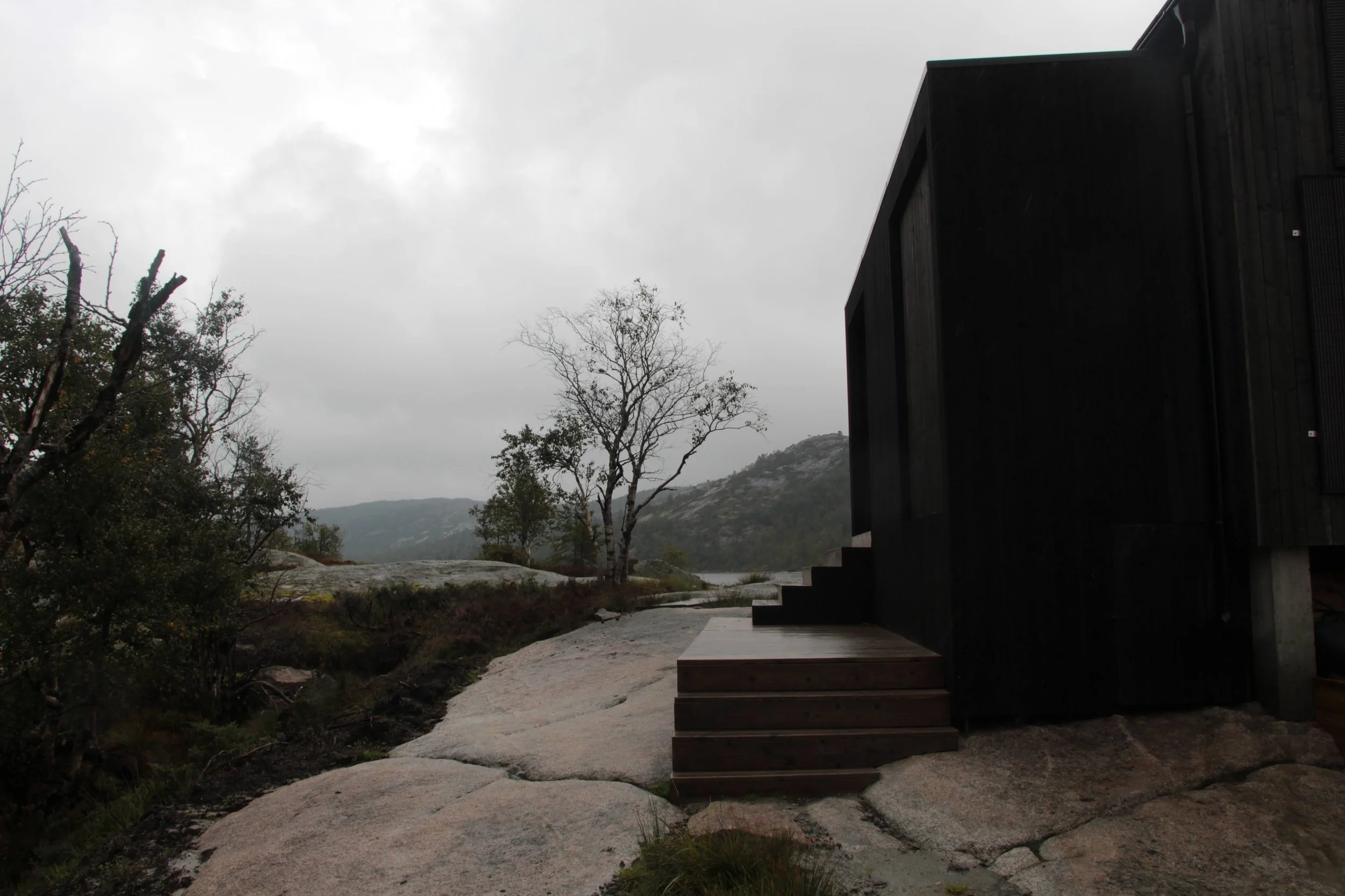 Fjellborg hytte - Et mørkt trehus med trapper som fører ut til et steinete uteområde, omgitt av trær, fjell og en skyet himmel i bakgrunnen.