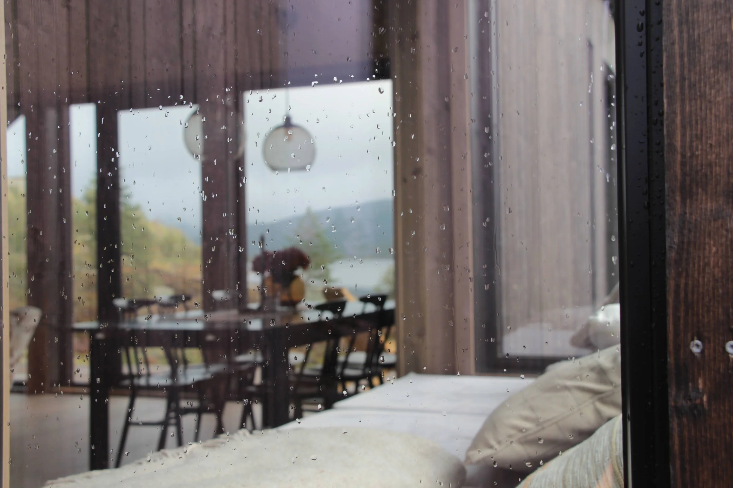 Fjellborg hytte interiør - Utsikt på en regnværsdag gjennom et vindu med regndråper, som viser en spisestue med et bord, stoler og en blomsterbukett utenfor.