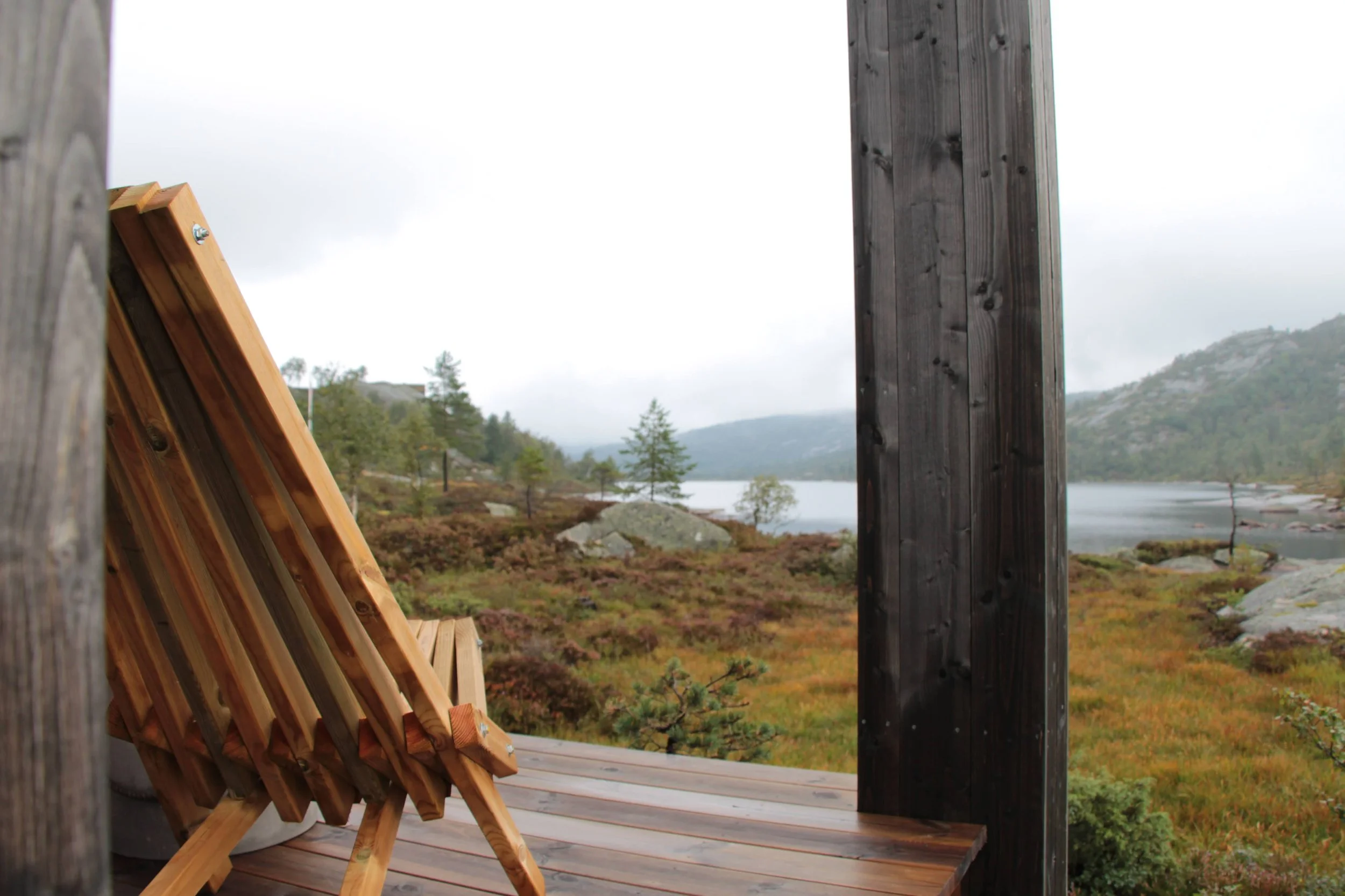 Fjellborg hytte - En treterrasse med en liggestol i tre som vender mot en innsjø omgitt av fjell og trær på en overskyet dag.