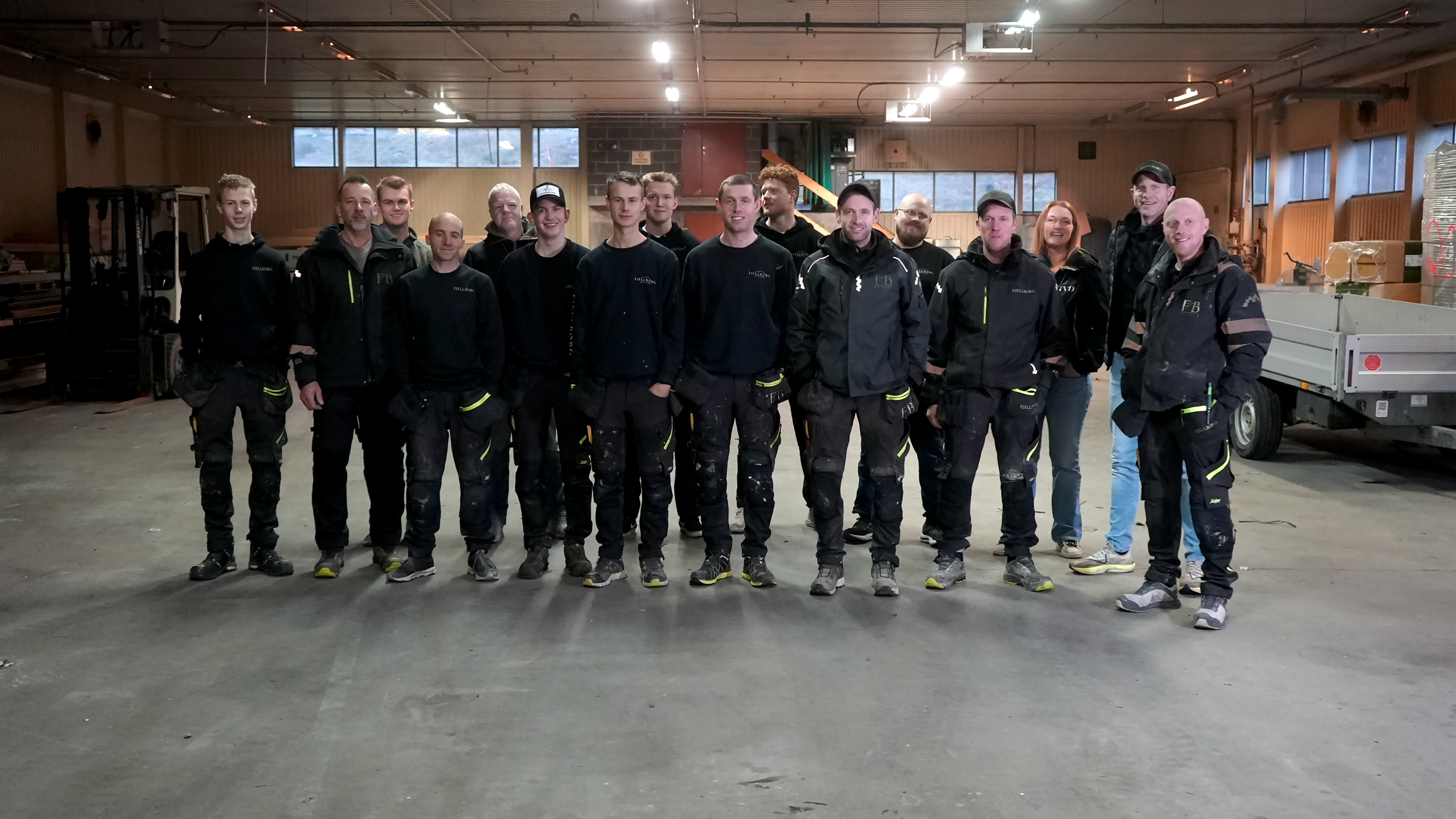 Fjellborg Hytter - En gruppe mennesker står sammen innendørs, mange av dem er kledd i svarte arbeidstøy med refleksdetaljer.