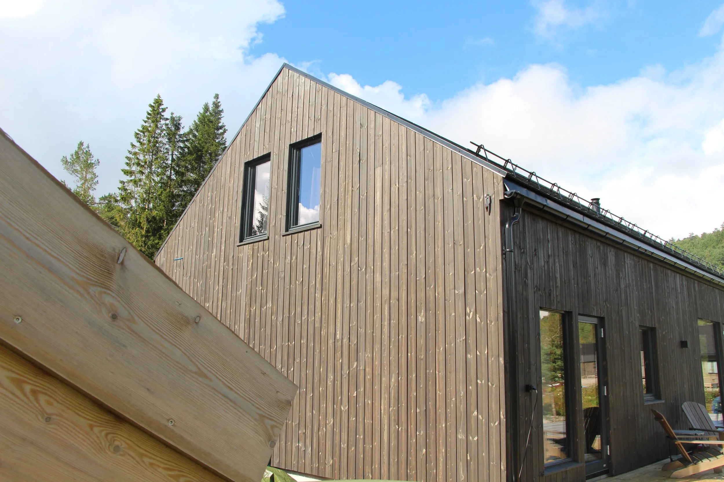 A modern wooden house with vertical planks, three large windows on the upper level, and a sliding glass door on the lower level, set against a background of trees and a partly cloudy sky.