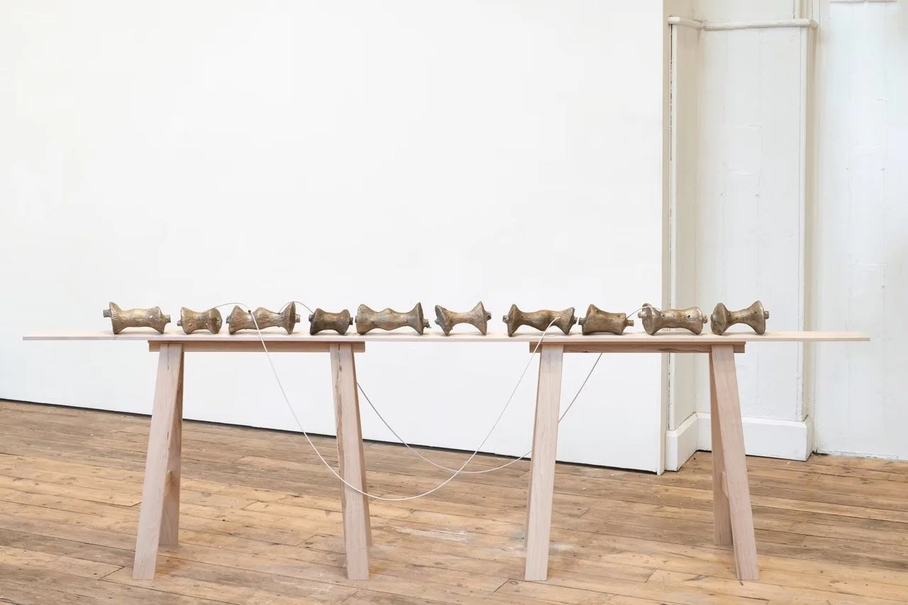 An art installation featuring eleven metallic dog sculptures connected with white strings, displayed on a long wooden table in a gallery with wooden floors and white walls.