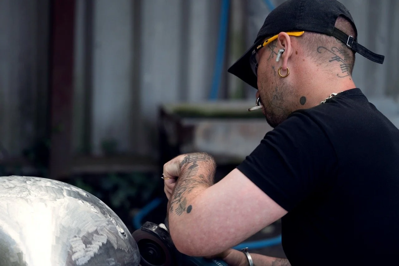 A man with tattoos and earrings wearing sunglasses and a backwards cap, working on a metallic object with a power tool.