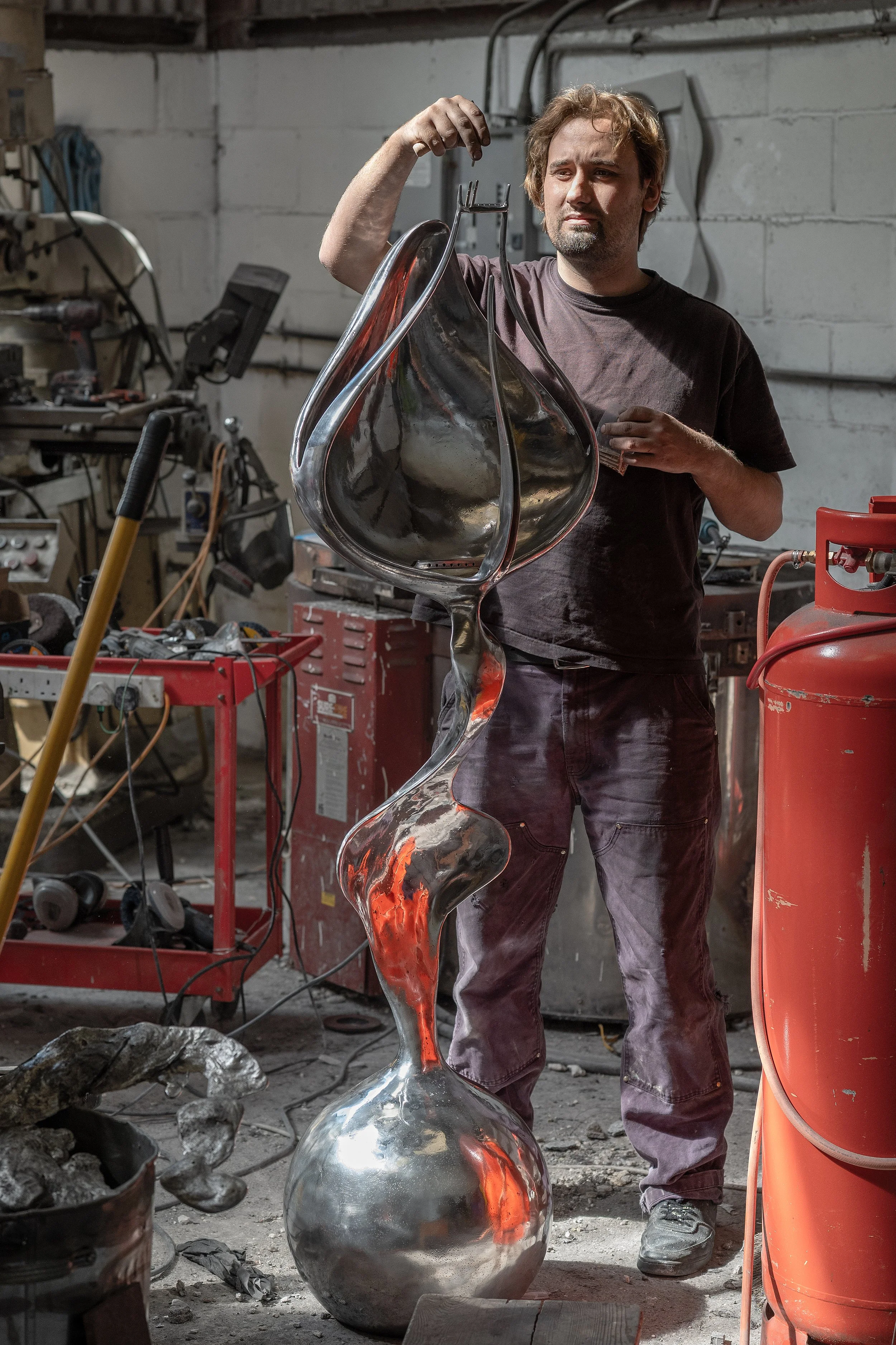 A man in a workshop holding a large, artistically curved metallic sculpture.