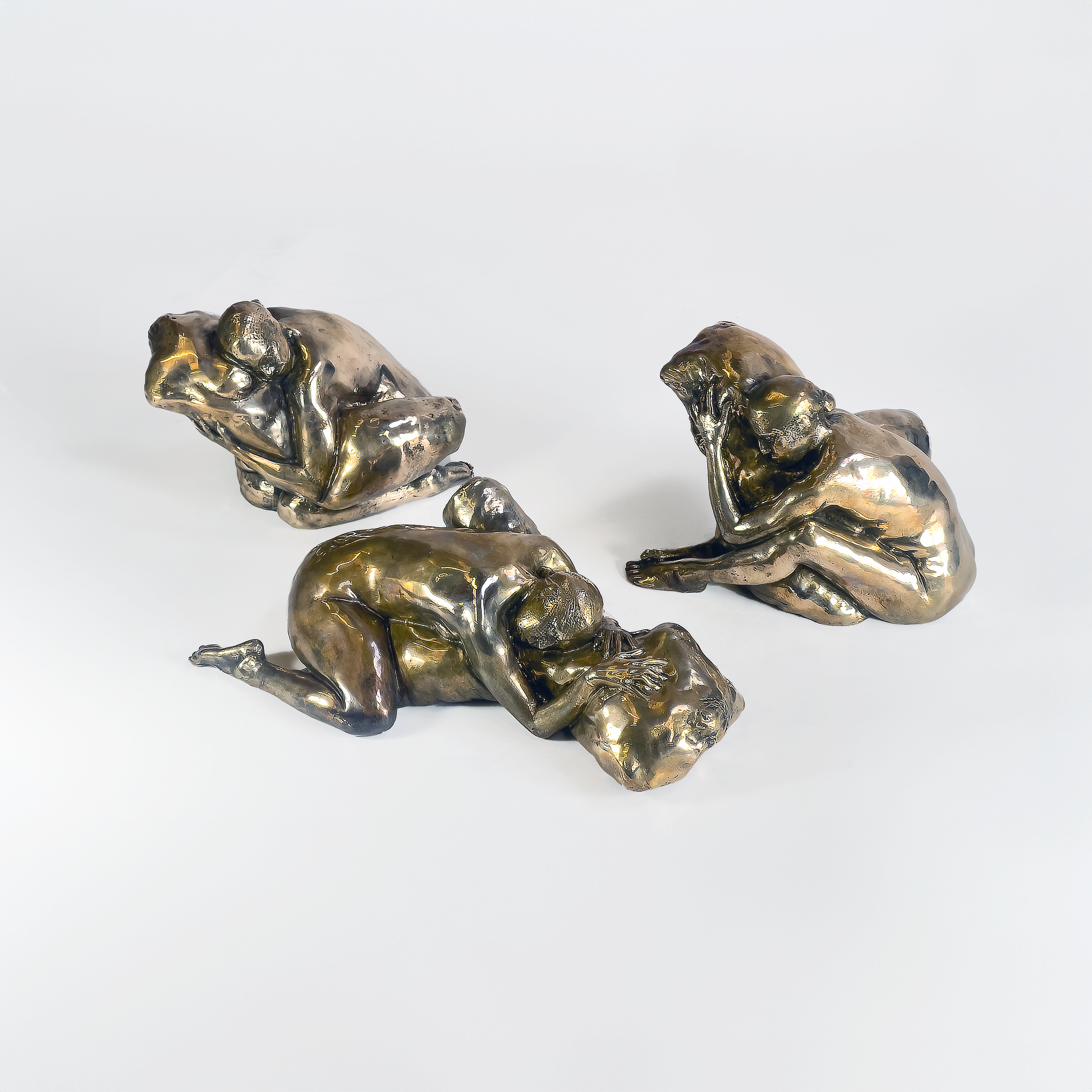 Three metallic sculptures of human figures in various seated and lying postures on a white background.