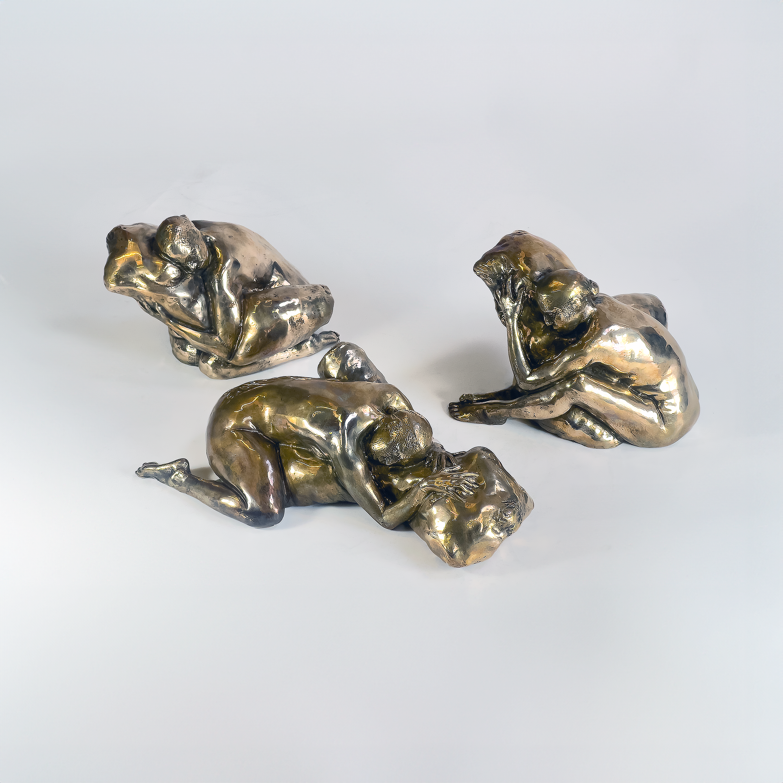 Three metallic sculpture figures of women in various seated and lying poses on a white background.