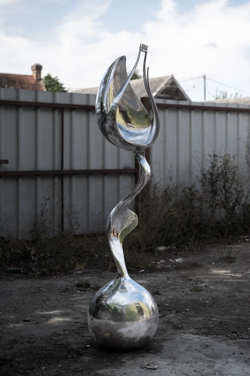 Silver abstract sculpture resembling a twisted elongated form with a fork at the top, placed outdoors on a dirt surface with a fence and rooftops in the background.