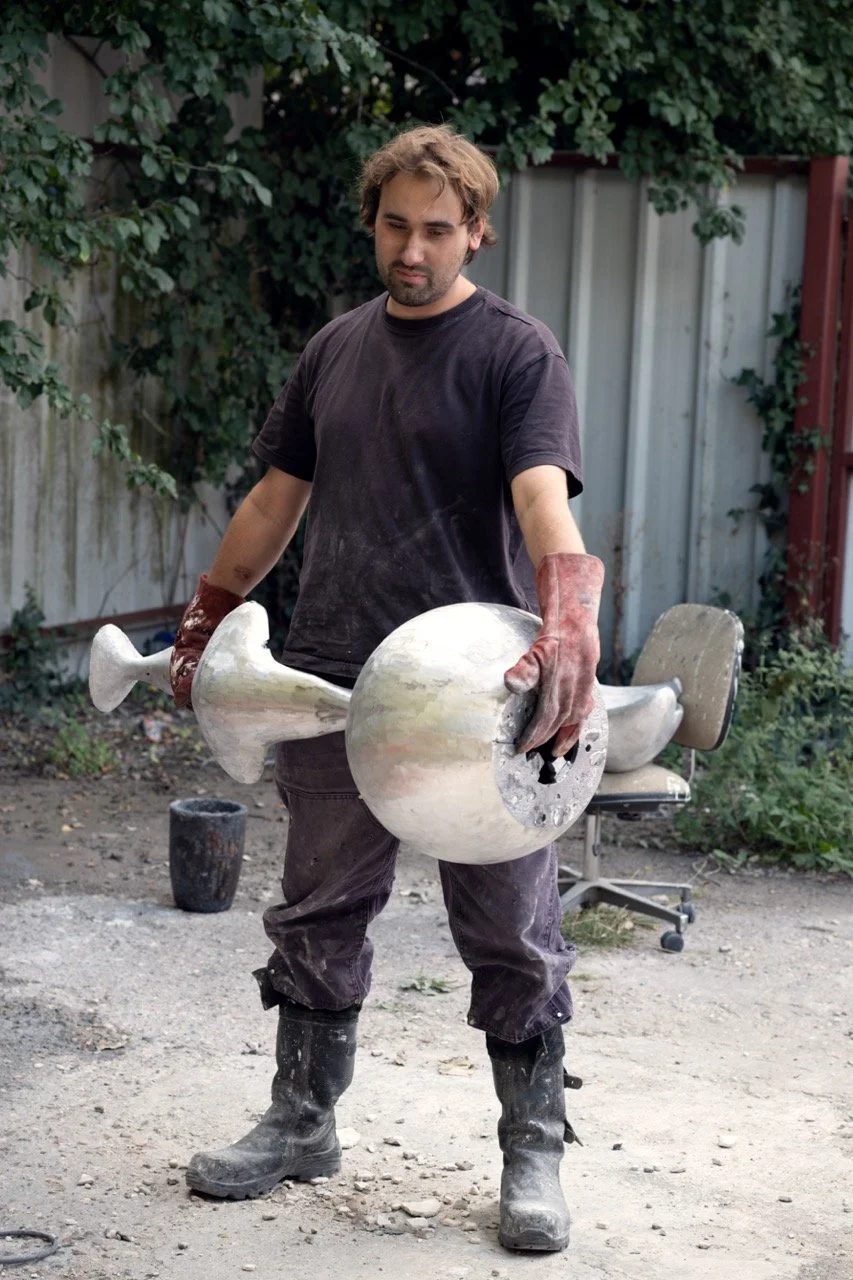 Man wearing black t-shirt, gloves, and boots holding a large metallic sculpture resembling a rocket in an outdoor backyard setting.