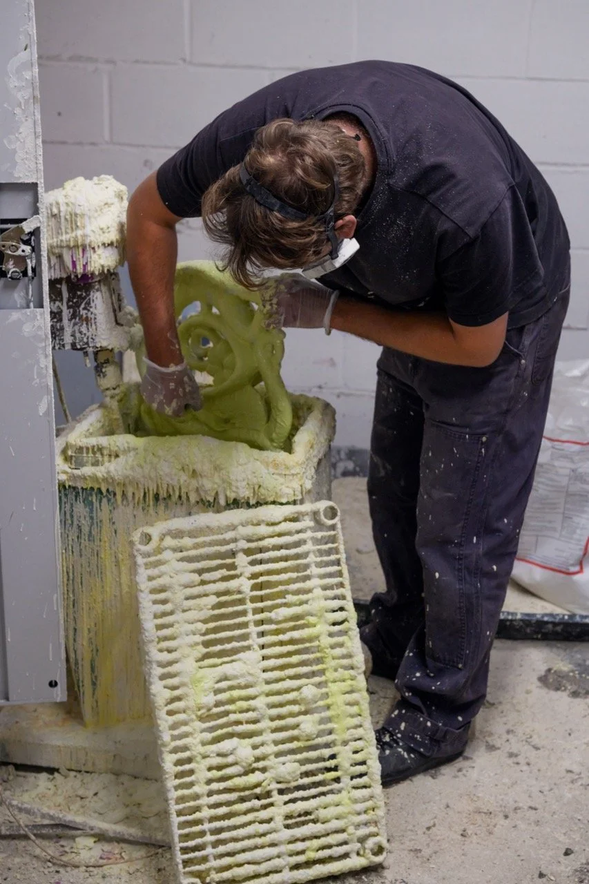 A person working on a foam sculpture with a grinder or similar tool, wearing gloves and safety glasses, in a workshop with a cinder block wall.