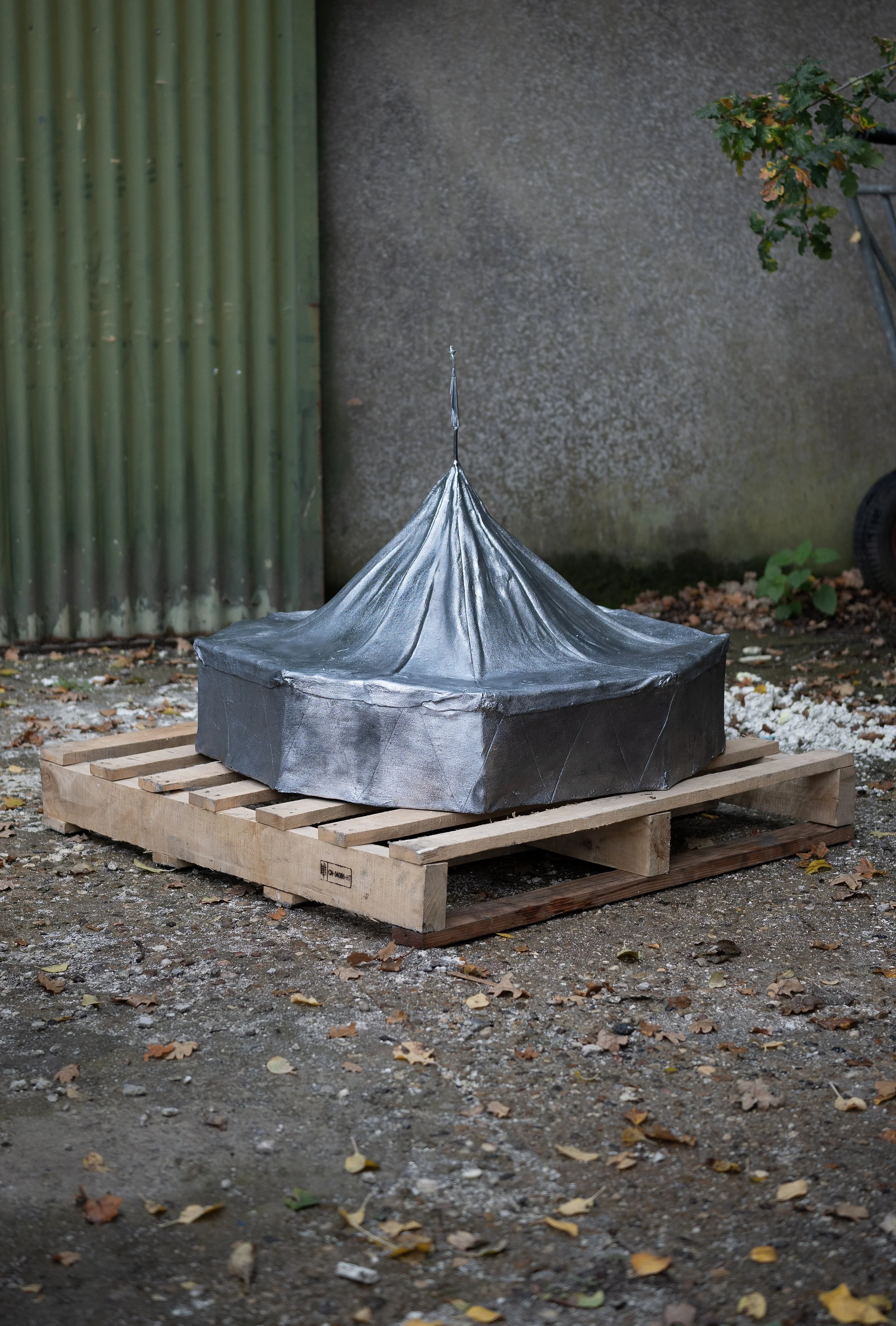 A metallic tent-like structure placed on a wooden pallet outdoors, surrounded by fallen leaves and a gravel ground.