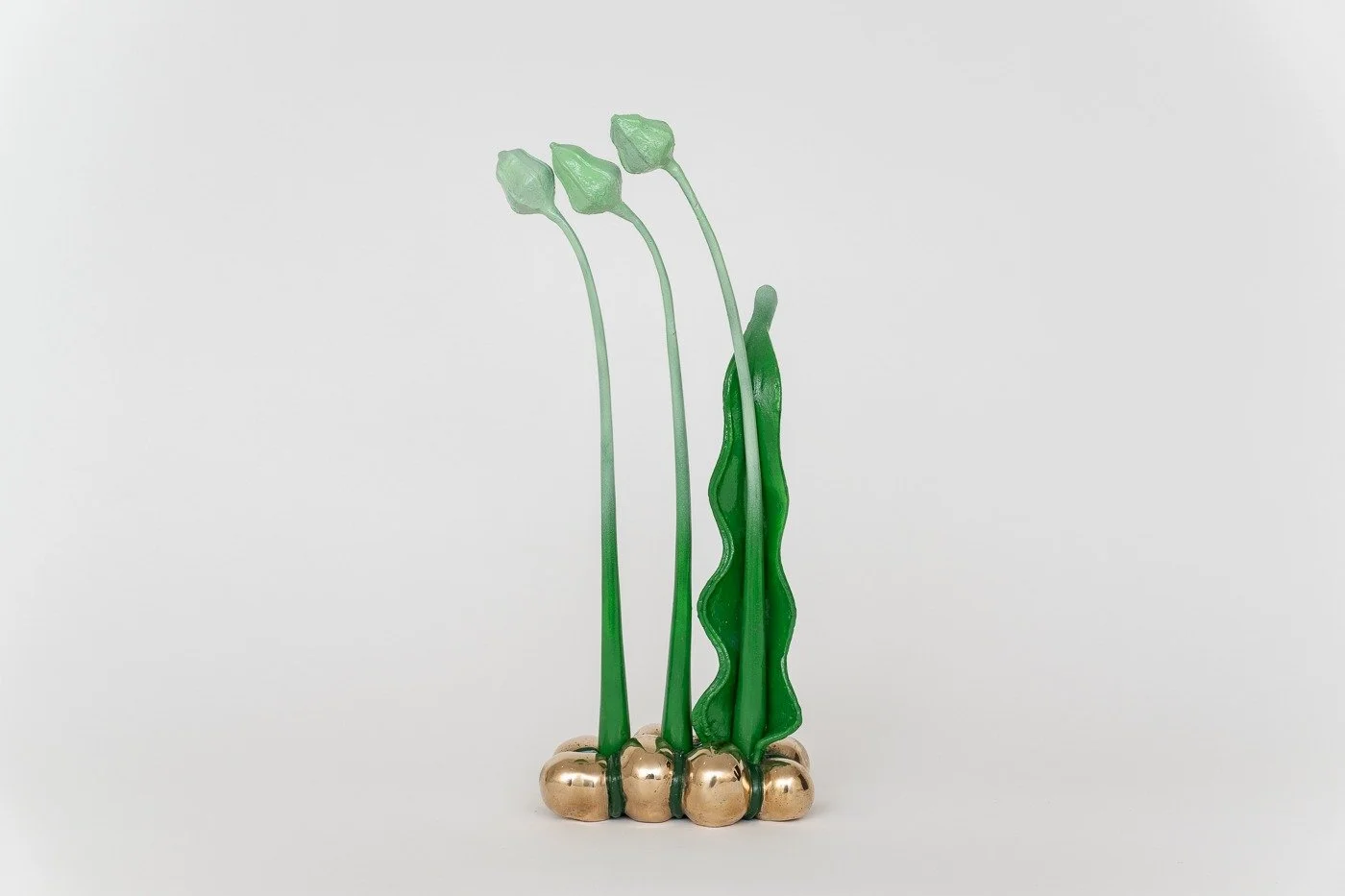 Contemporary glass sculpture resembling plant stalks with three green flower buds and a wavy green leaf, mounted on a gold-colored base, on a plain white background.