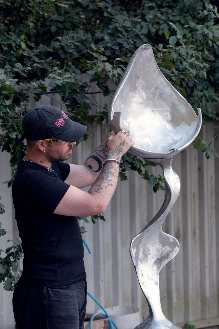 Man in black cap and sunglasses working on a large metallic sculpture resembling a spoon with a twisted handle, outdoors with trees and a wooden fence in the background.