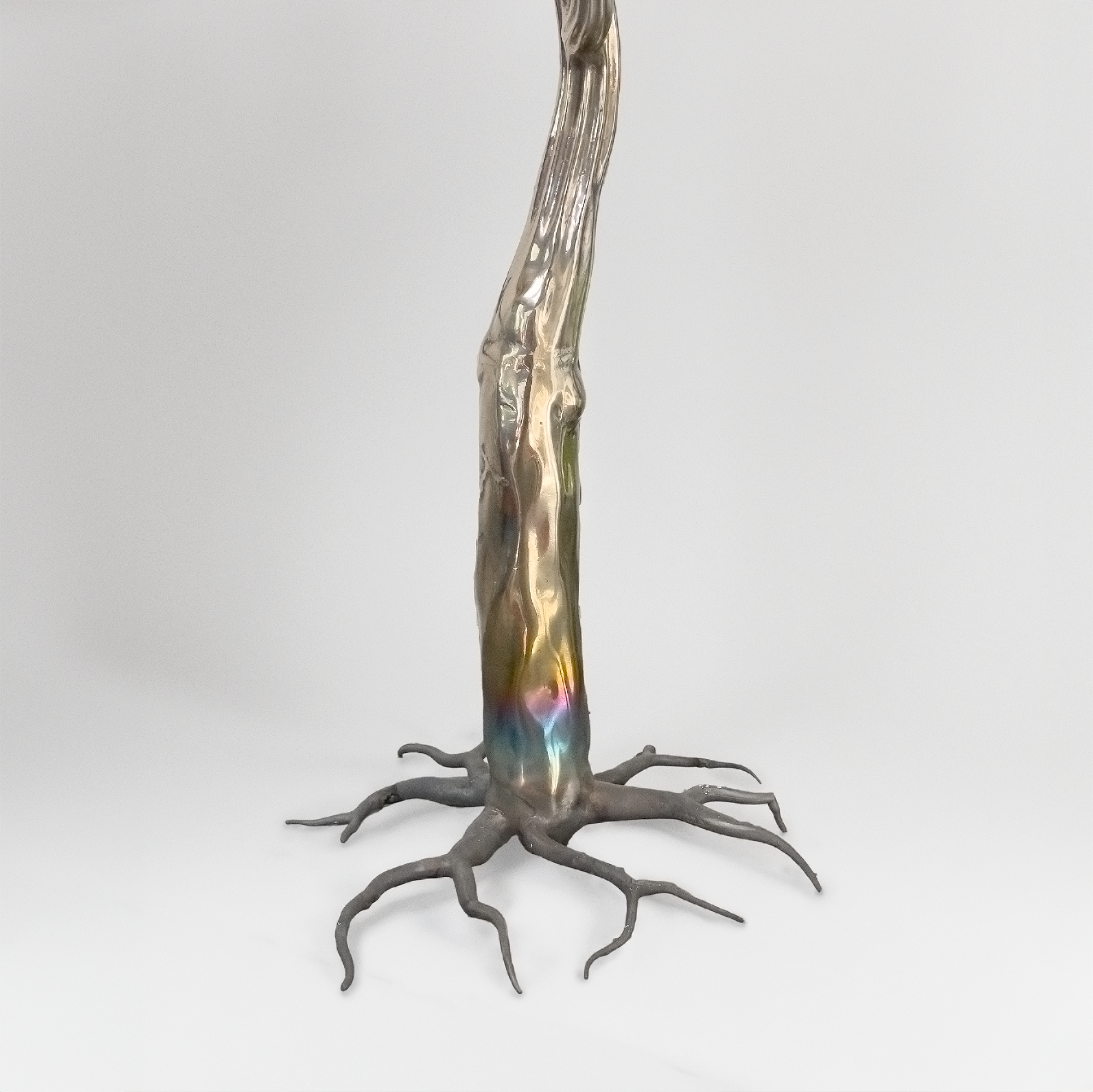 A metallic sculpture resembling a tree with rainbow-colored reflections on its trunk and exposed roots at the base, set against a plain white background.